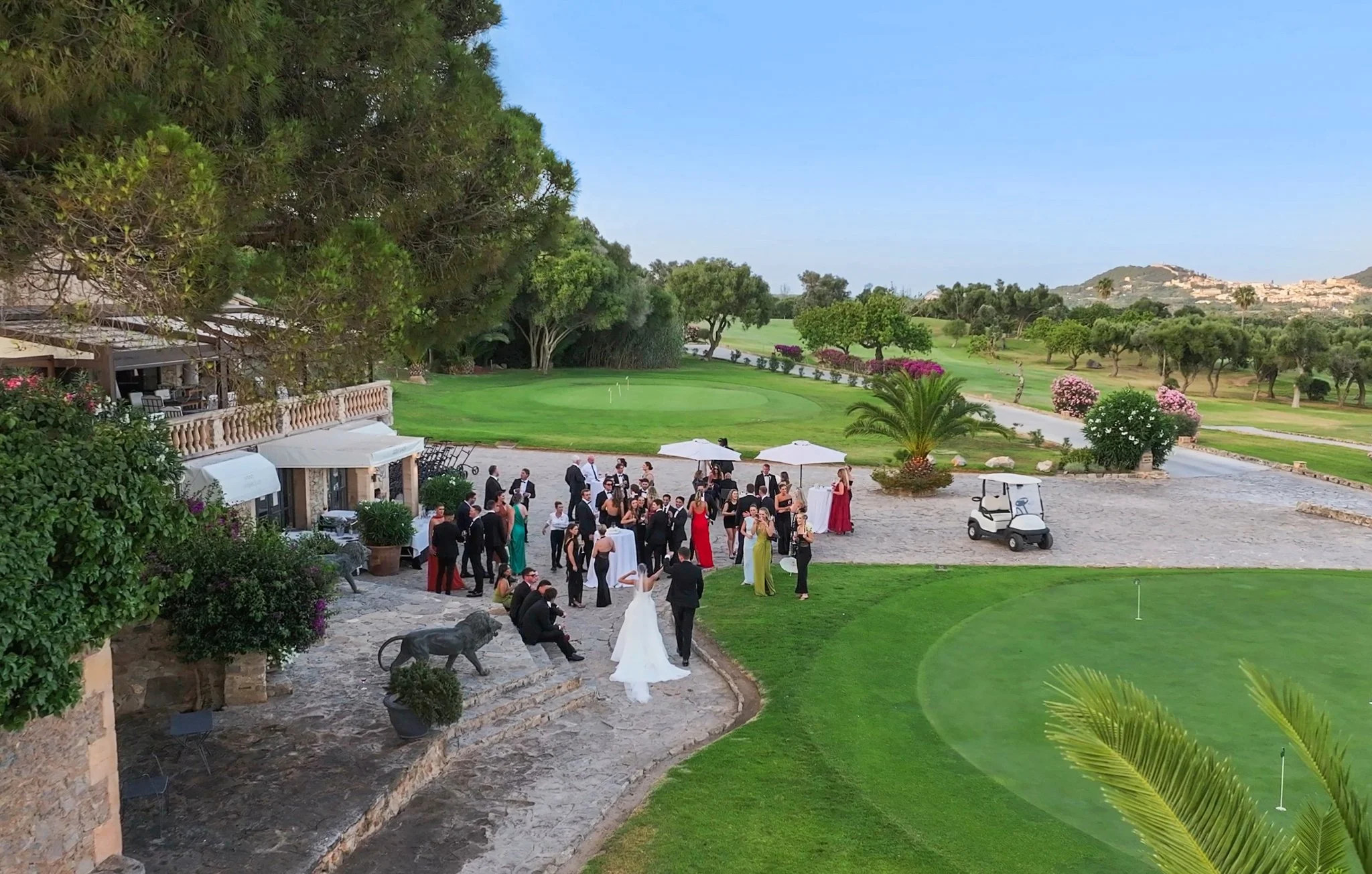 An outdoor event with people dressed formally, likely a wedding or celebration, near a golf course with green grass, colorful bushes, trees, and hills in the background. There are umbrellas and a golf cart.