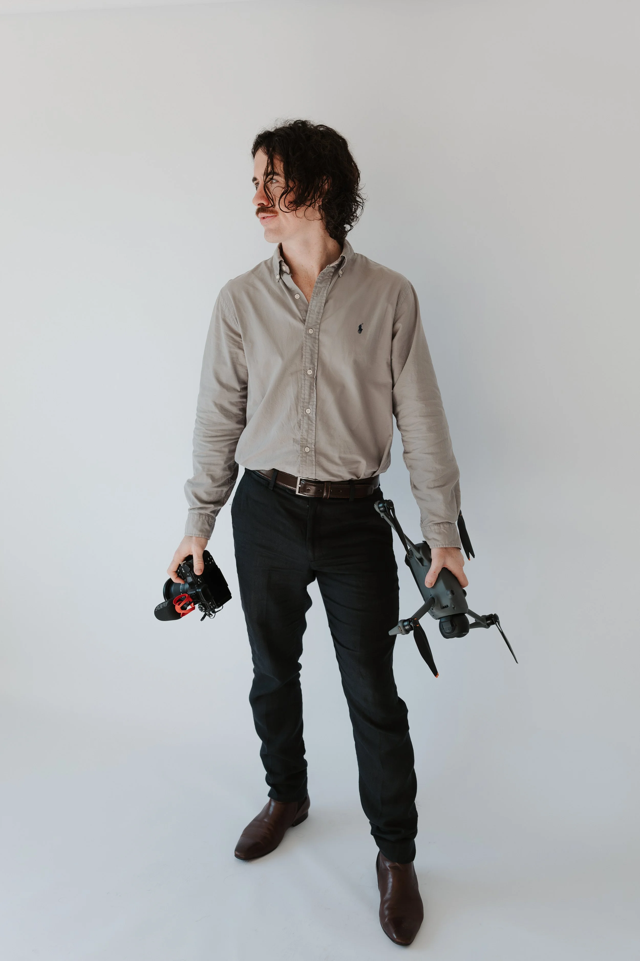 A man with curly dark hair, a mustache, and light skin wearing a beige button-up shirt, dark pants, and brown boots, holding a camera in his right hand and a drone in his left hand, standing against a plain white background.