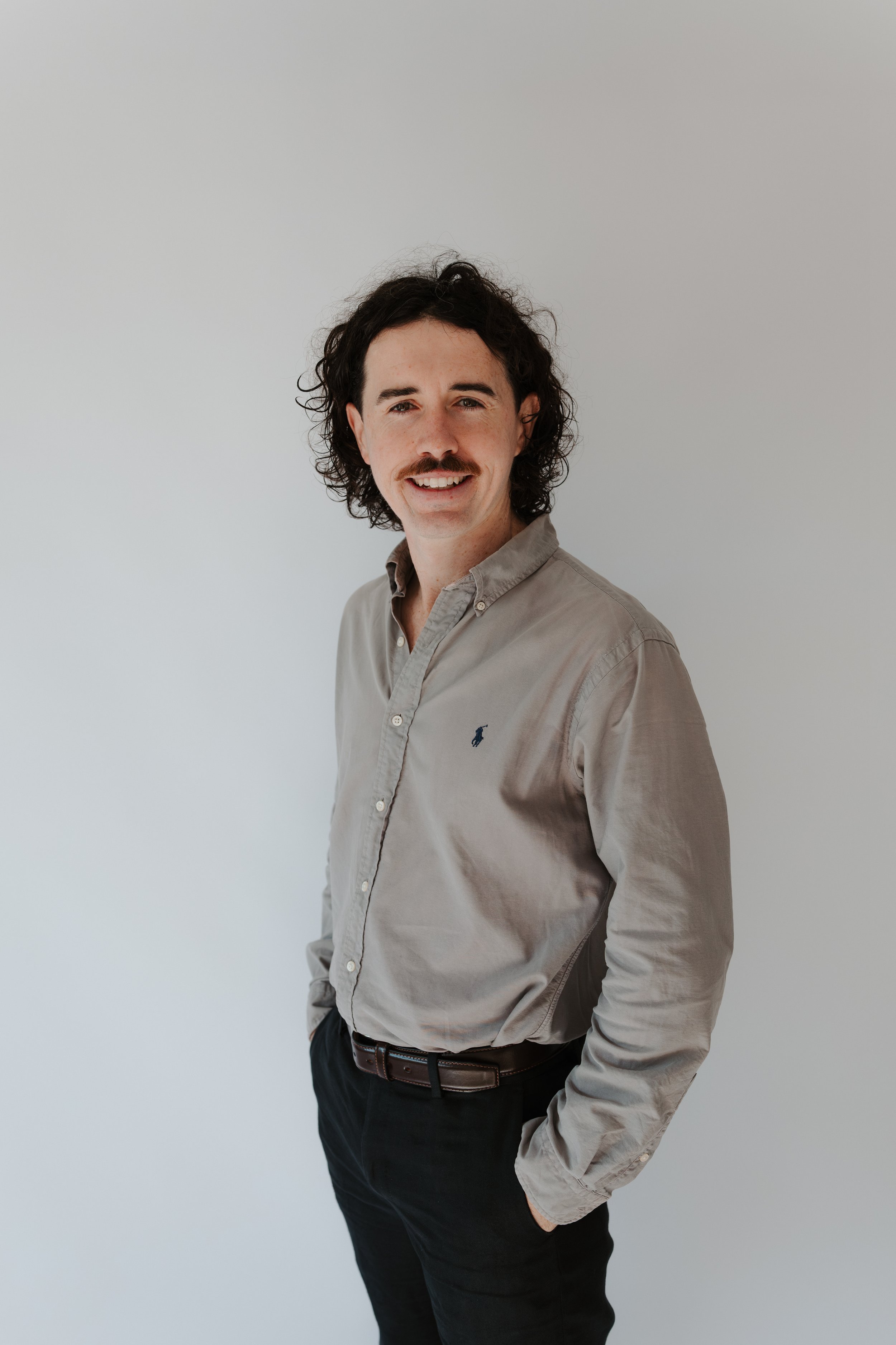 Portrait of a man with curly dark hair, mustache, wearing a beige button-up shirt and black pants, standing against a plain white background.