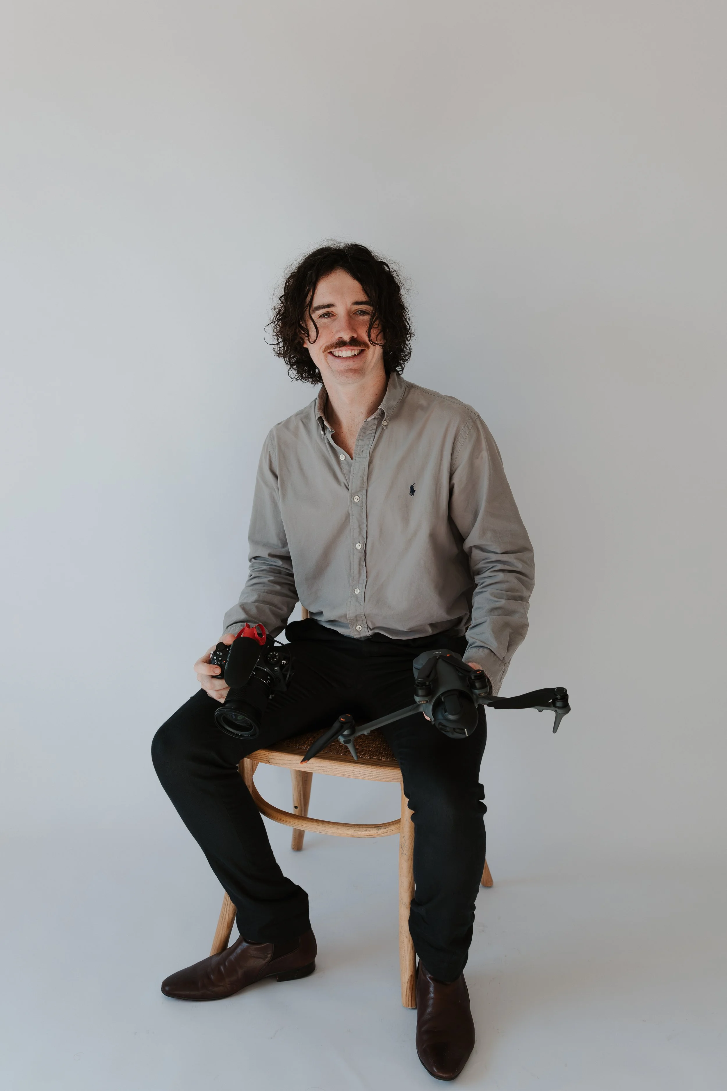 A man with dark curly hair and a mustache sitting on a wooden stool against a plain light gray background, holding a camera in his right hand and a drone on his lap, dressed in a beige button-up shirt and black pants with brown boots.