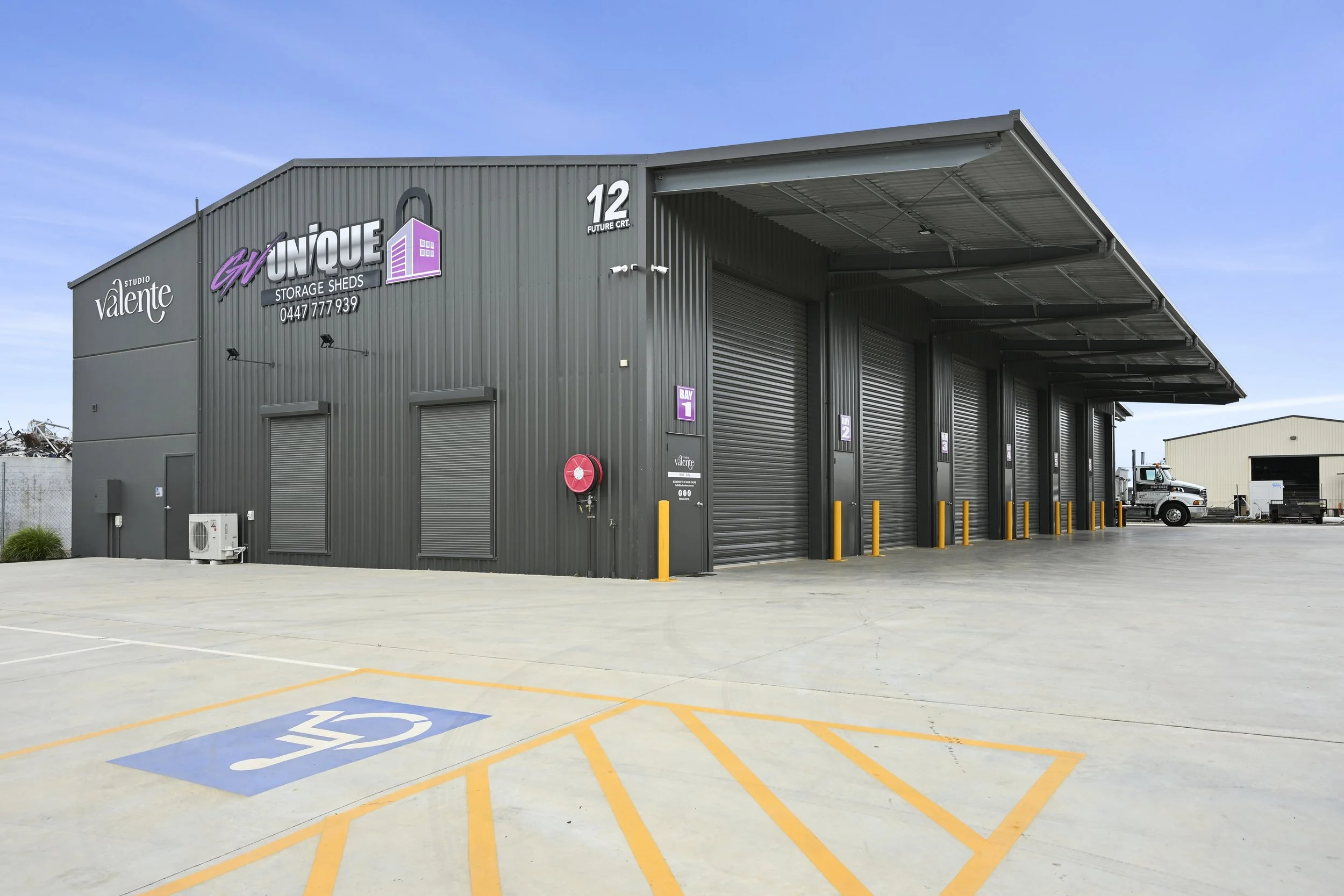 A large gray metal storage shed with multiple roll-up doors in a parking lot. There are yellow parking barriers in front of the doors and a handicapped parking space nearby. The building has signage for "GV UNIQUE Storage Sheds" with a purple logo and contact number. The sky is clear and blue.