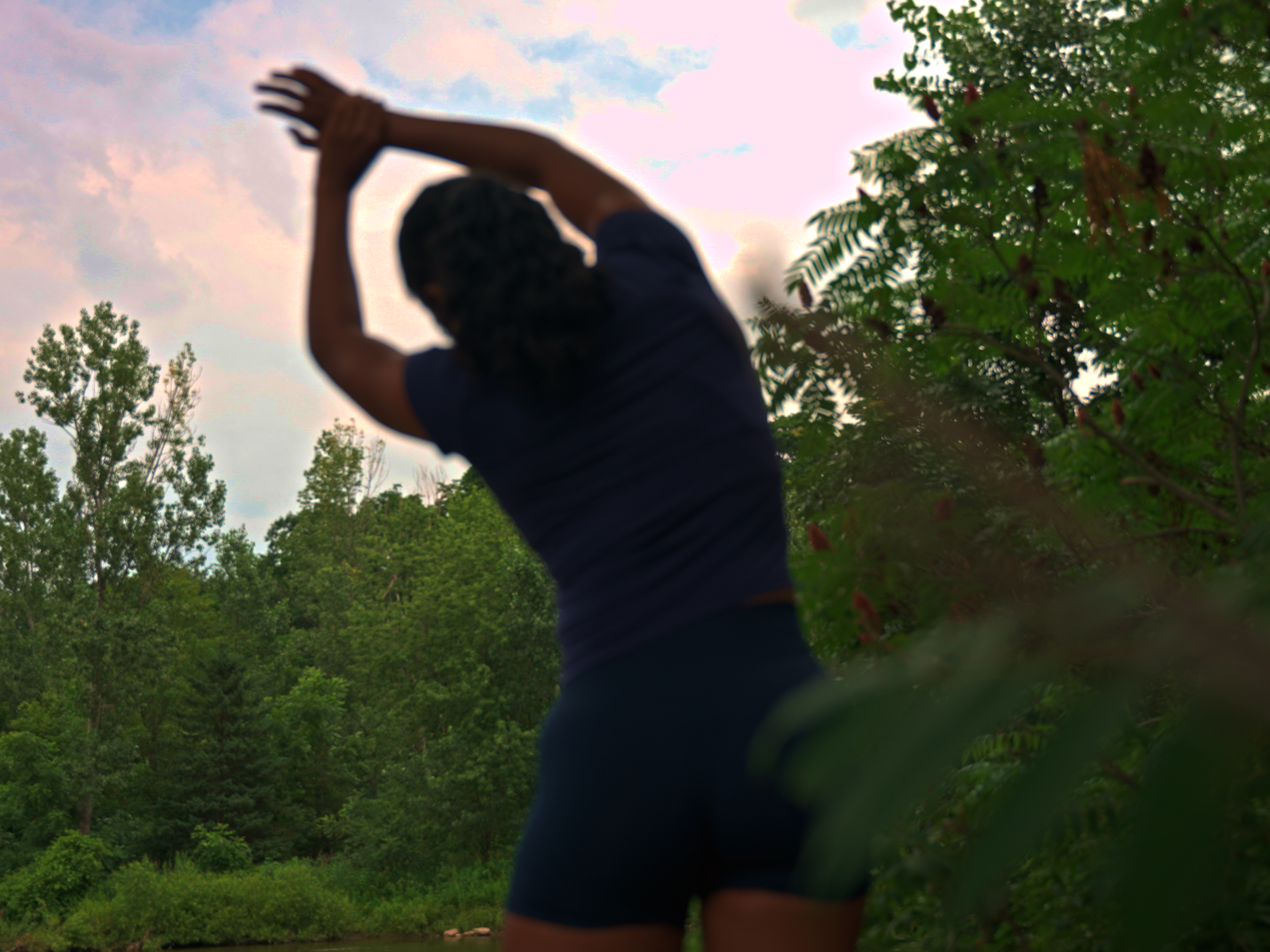 A person with dark hair tied in braids, wearing a dark shirt and jeans, raises their hands above their head in a natural outdoor setting with green trees and cloudy sky.