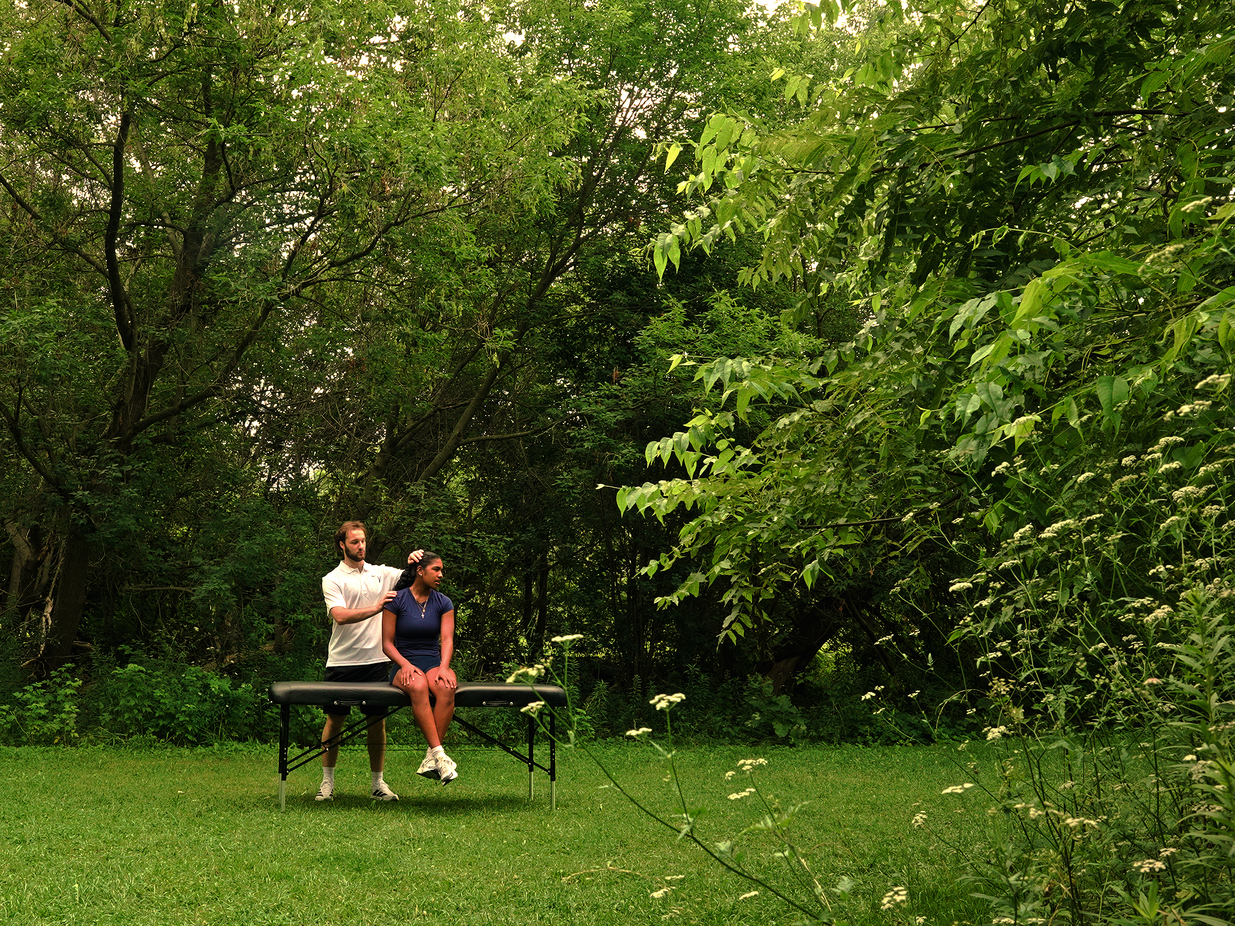 Two people, a man and a woman, are outdoors in a lush green wooded area. The man appears to be giving a massage or therapy to the woman, who is seated on a black table or bench. The woman is wearing a navy blue t-shirt and shorts, while the man is dressed in a light-colored shirt and dark shorts.
