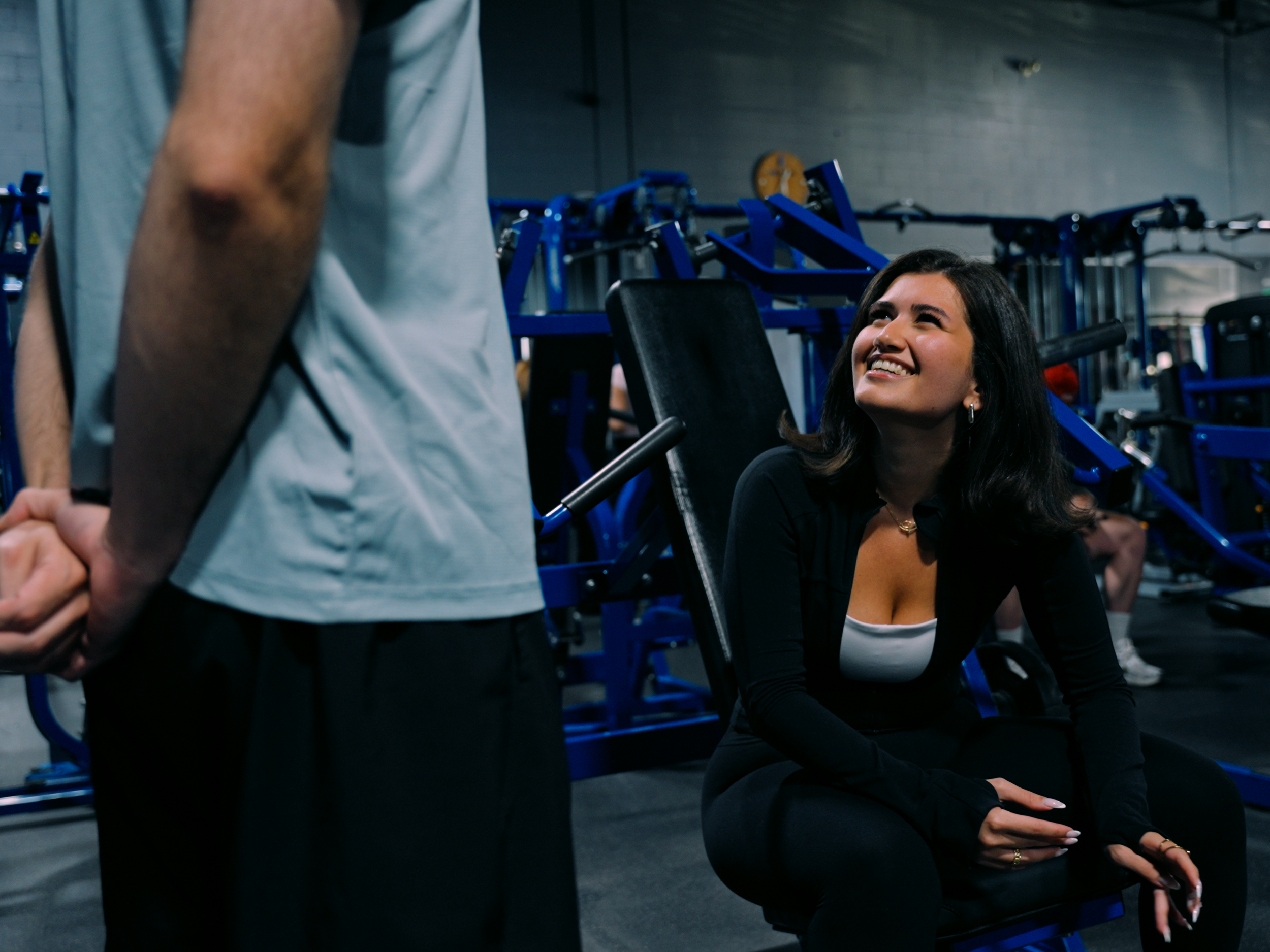 A woman with dark hair and earrings sitting on a workout bench in a gym, looking up and smiling at a person standing in front of her, with gym equipment in the background.