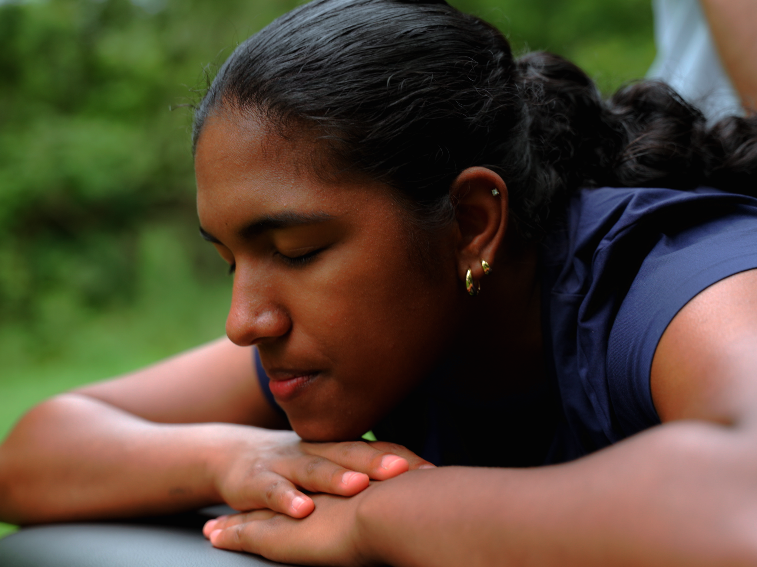 A young girl with dark hair and earrings rests her head on her folded arms outdoors on a cloudy day, with a green blurred background.