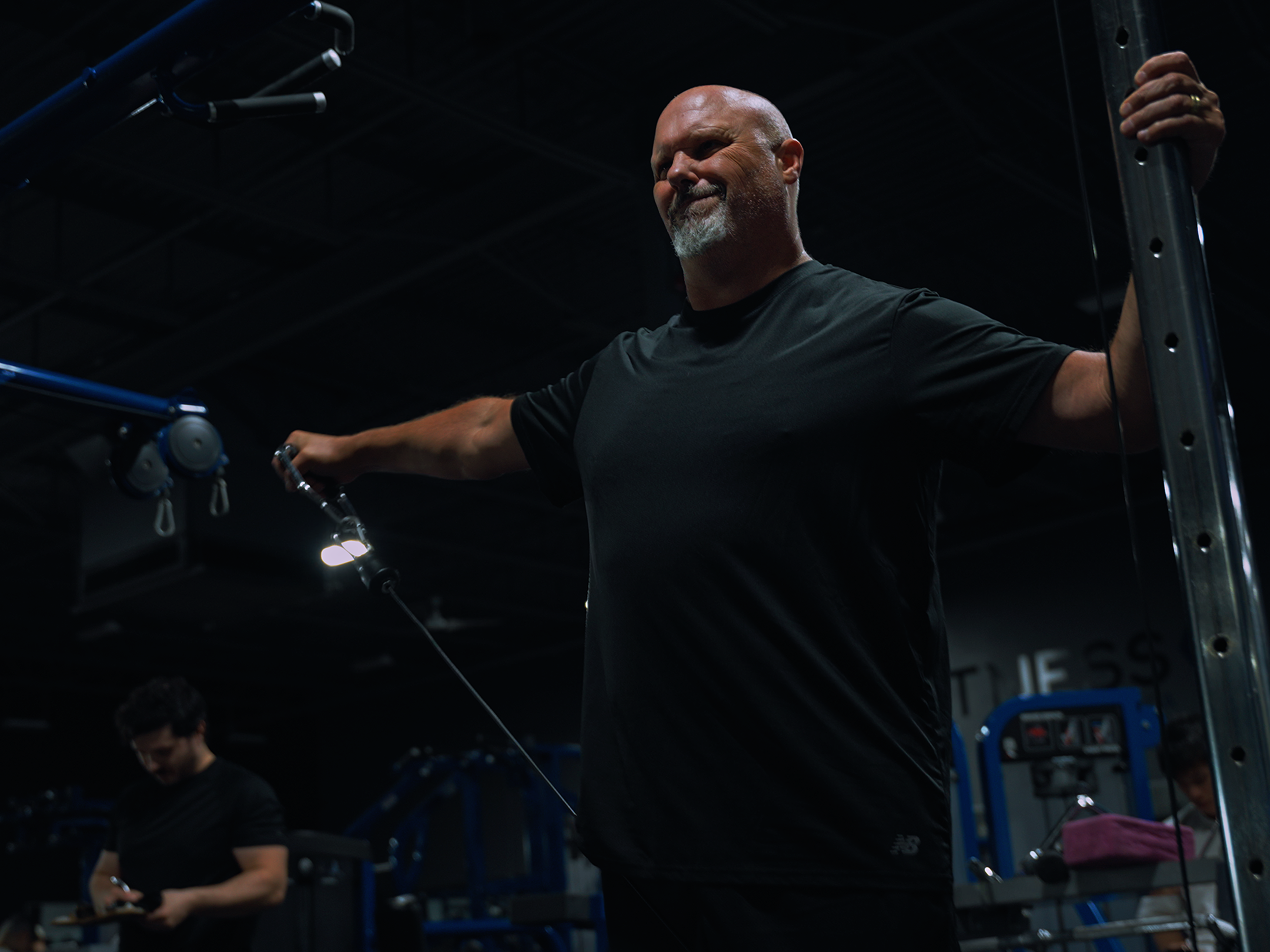 A man with a shaved head and beard smiling, wearing a black shirt, standing in a dark indoor gym environment with fitness equipment around him.