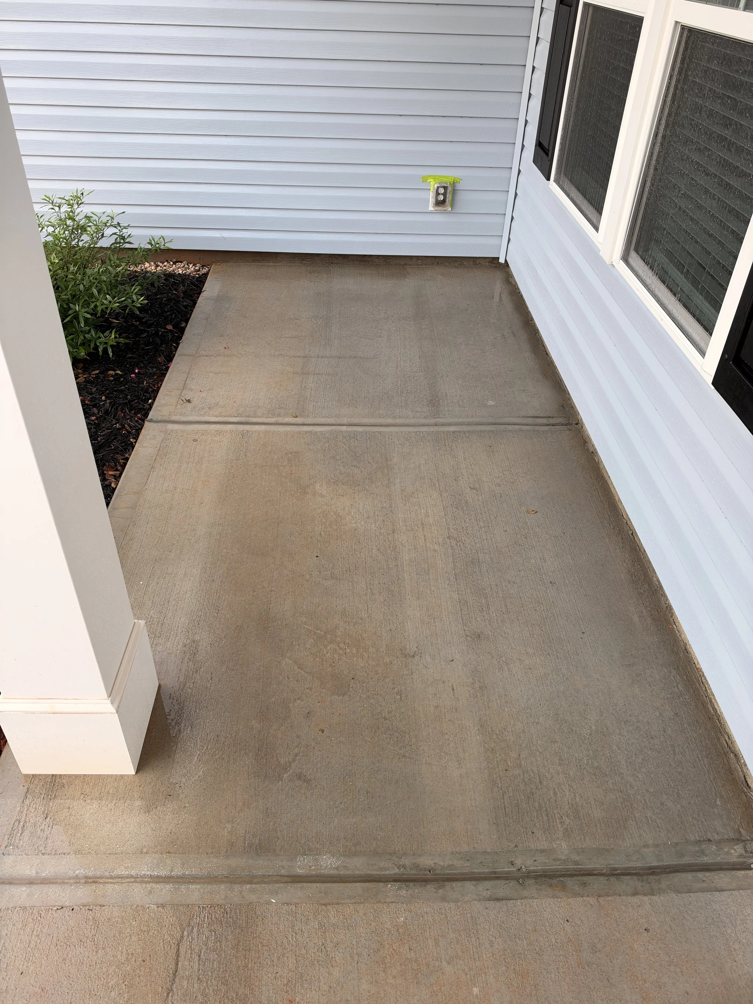 Concrete front porch beside light blue house siding, screened door, exterior outlet, and landscaped garden bed