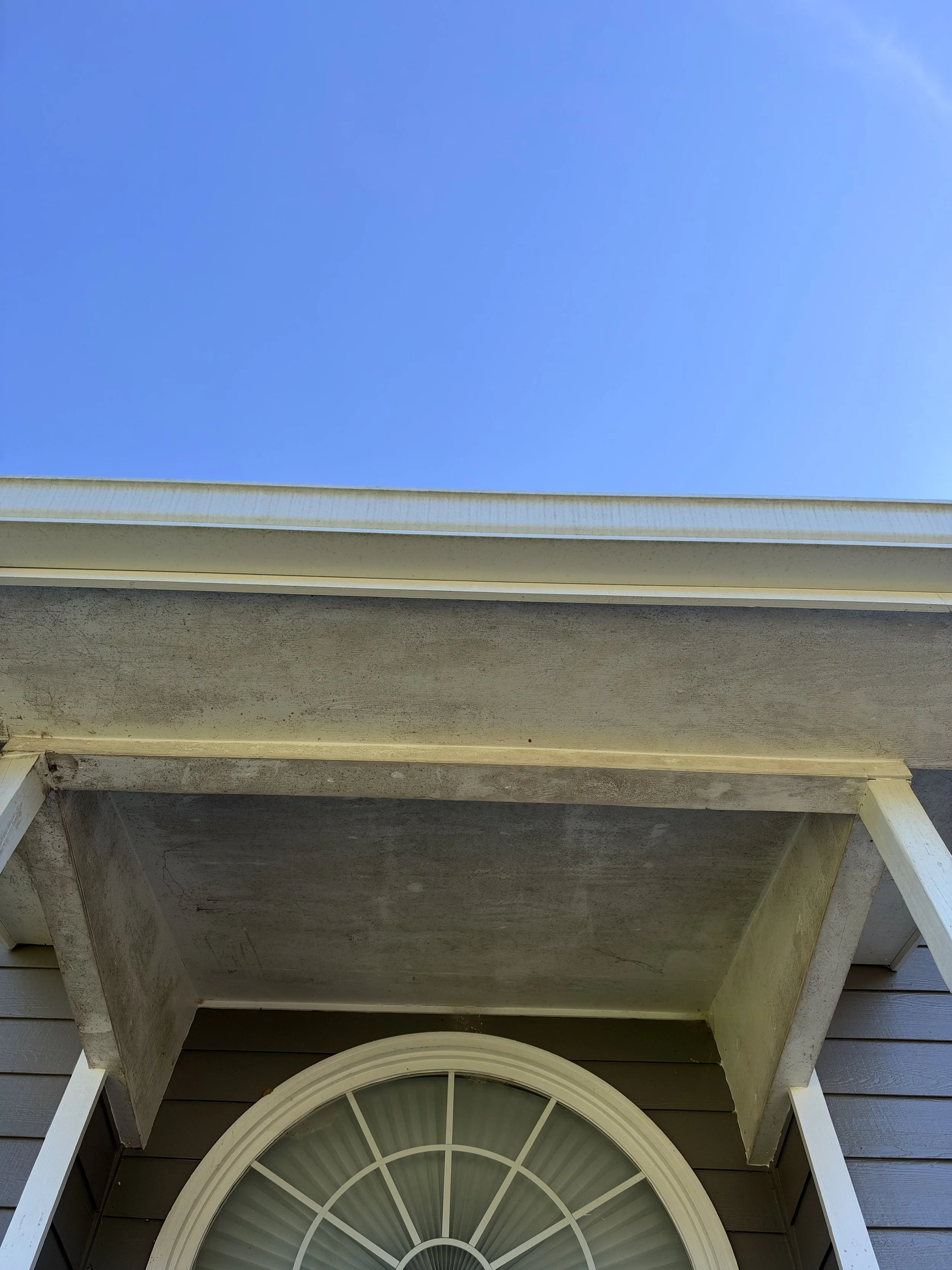 Close-up of a house exterior with blue sky, weathered gable trim, and a semi-circular decorative window.