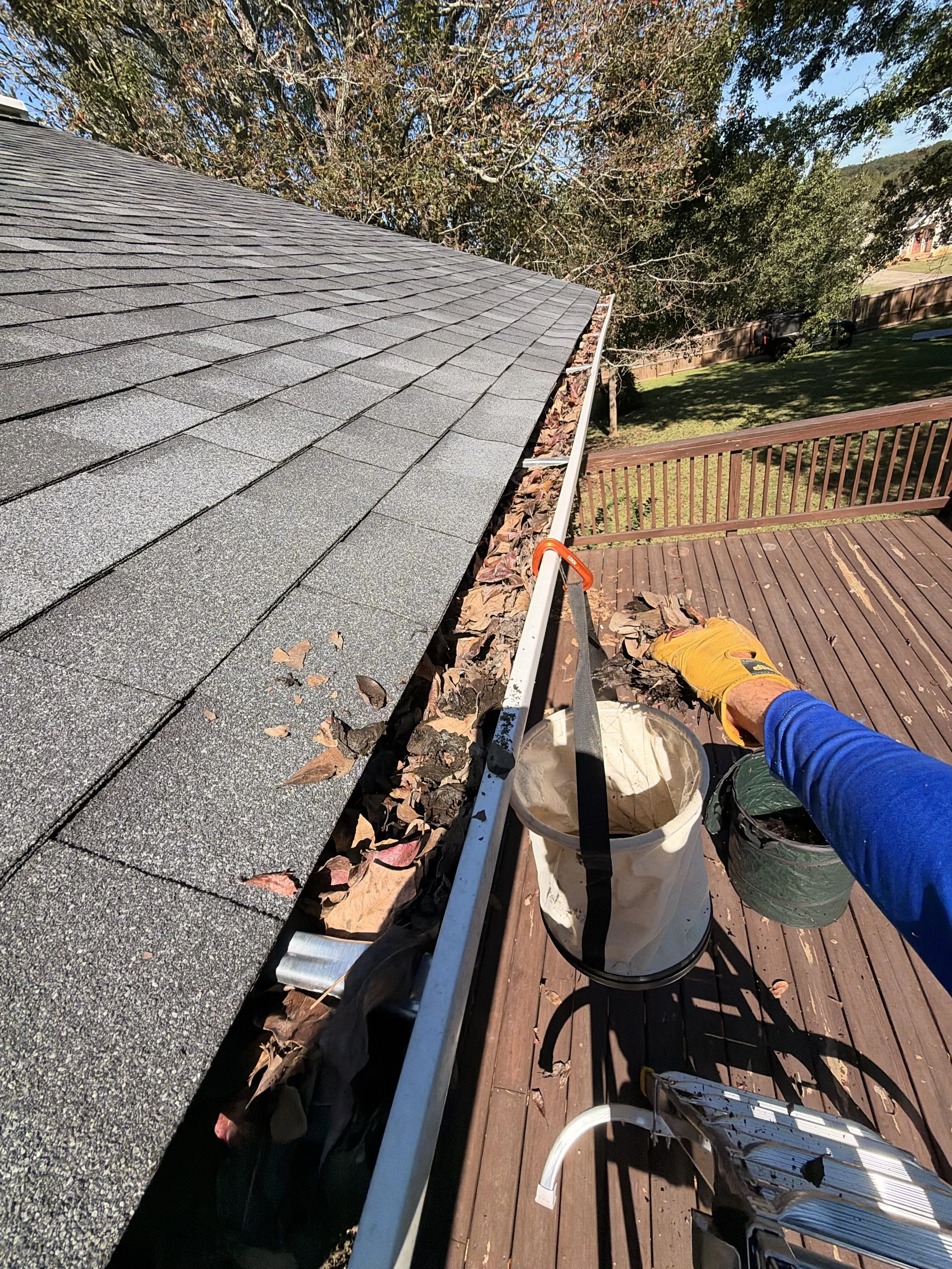 Roof worker clearing leaves from a gutter beside gray asphalt shingles and a wooden deck.
