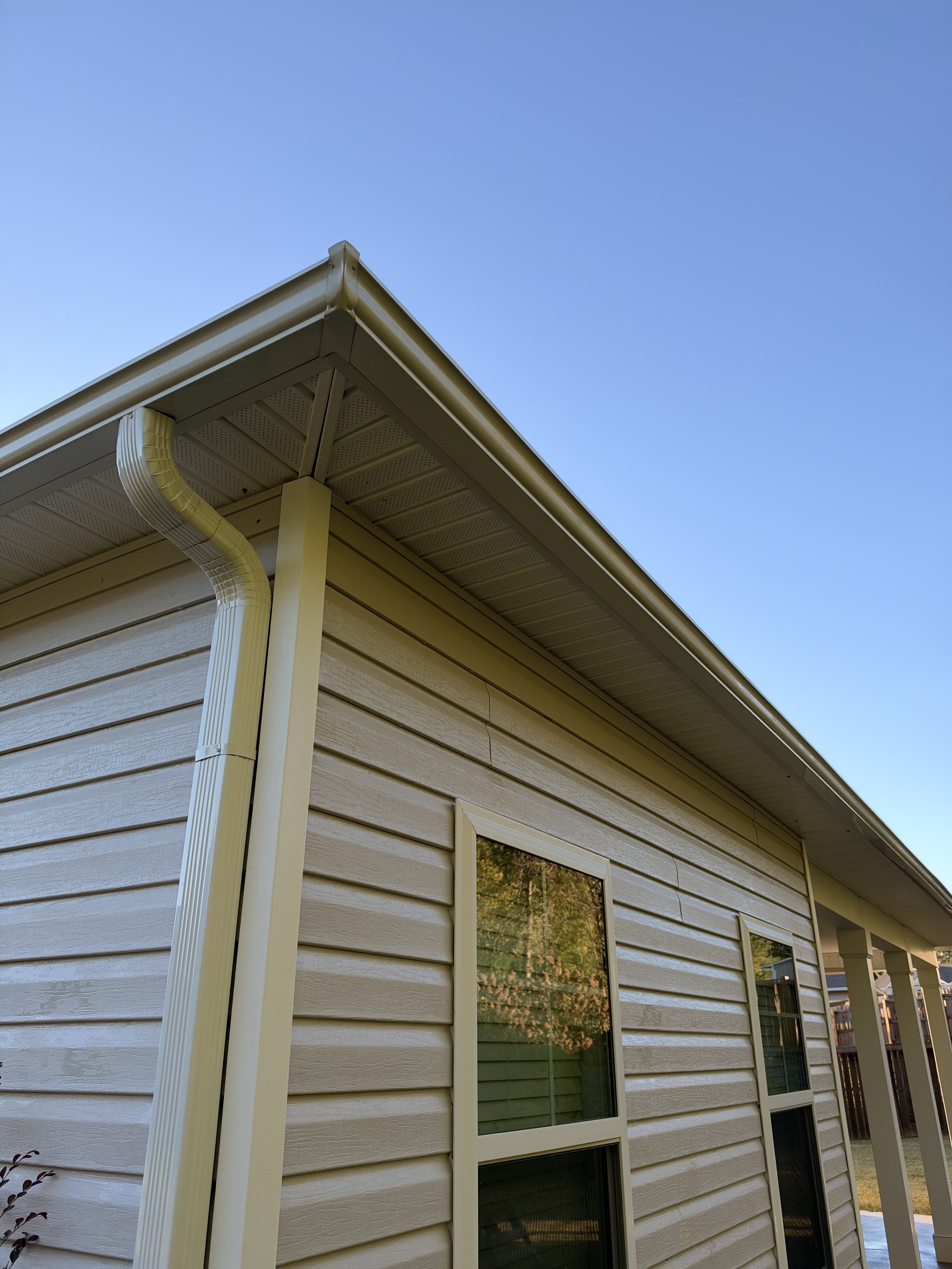 Close-up view of a house exterior with light siding, gabled roof, gutters, downspout, windows, and blue sky
