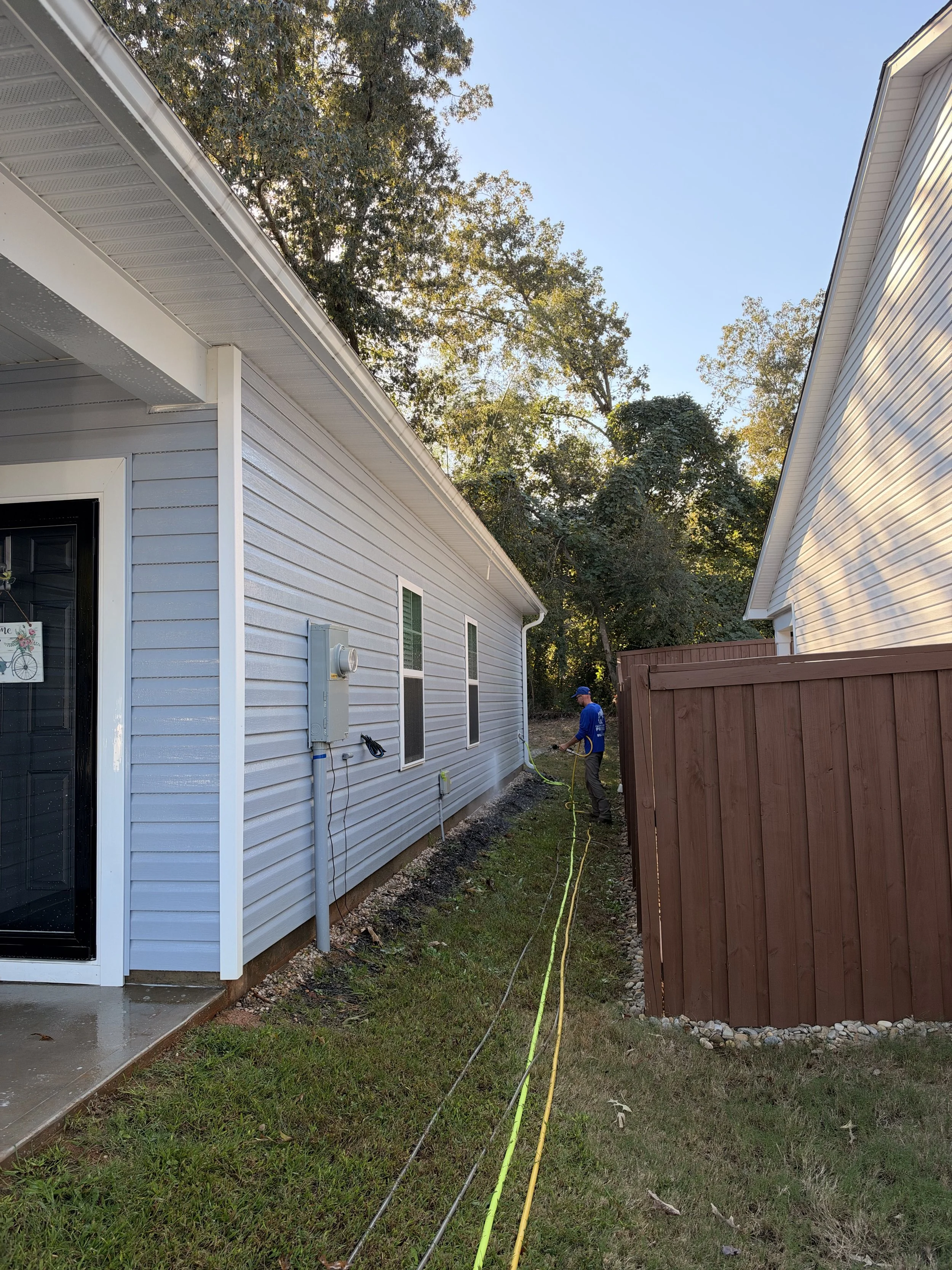 Worker pressure-washing the side of a gray vinyl-sided house beside a brown privacy fence.