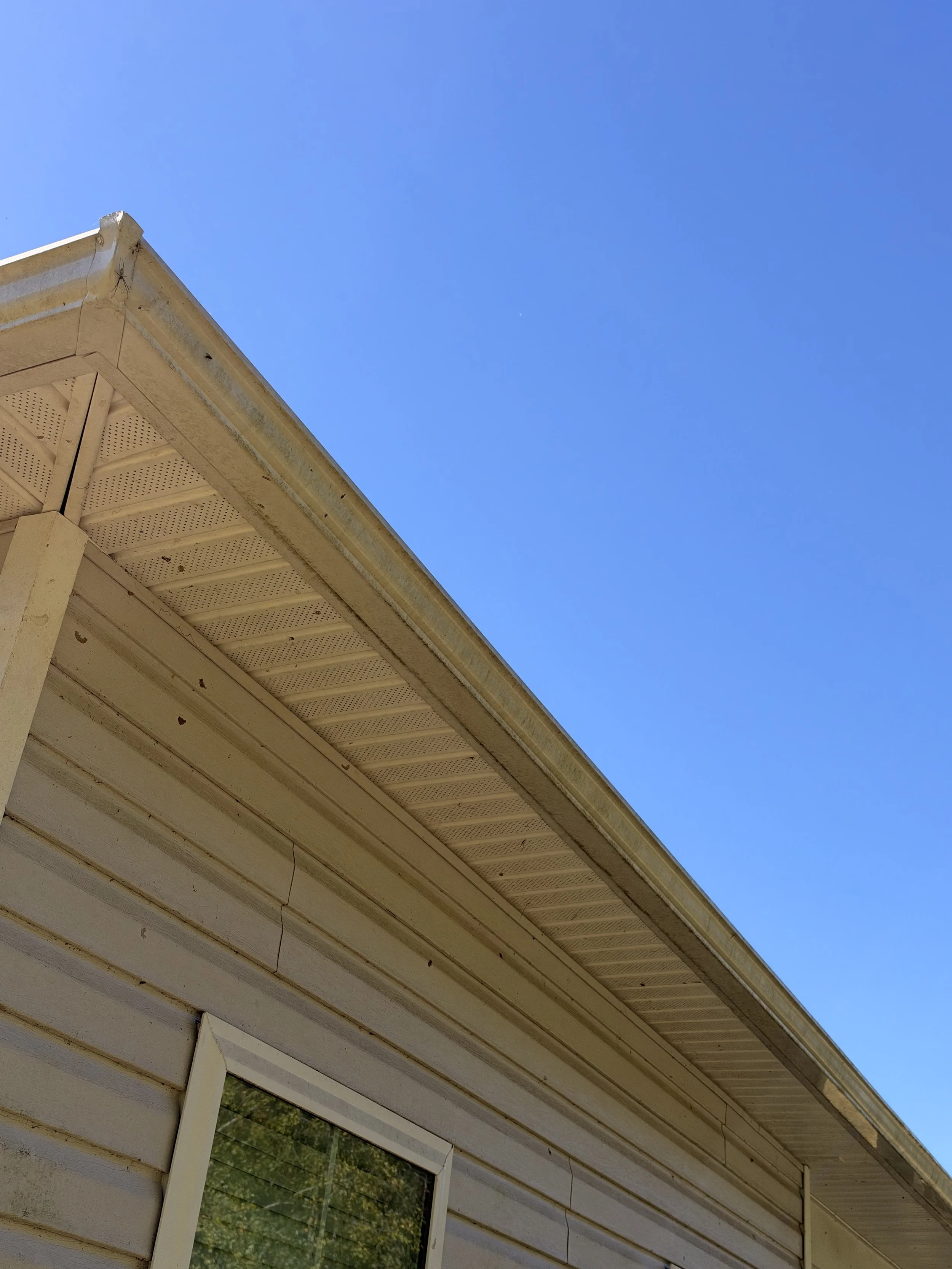 Upward view of a weathered white house roof eave with perforated soffit against a clear blue sky