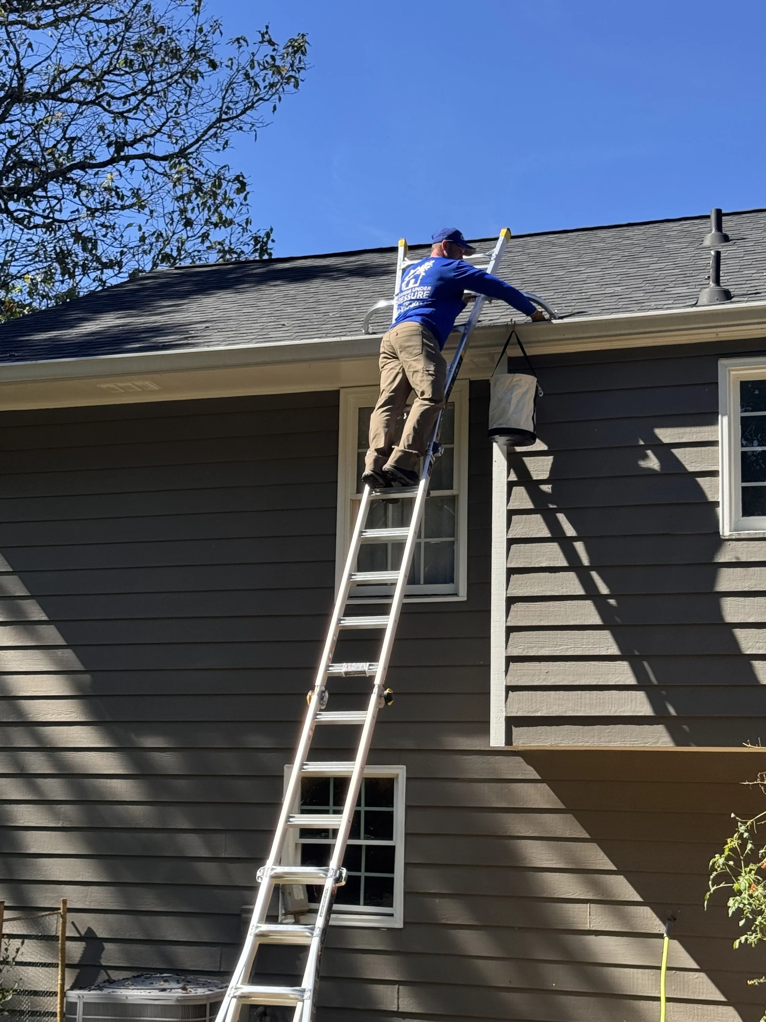 Worker climbing an extension ladder beside a house roof and gutter under a clear blue sky.
