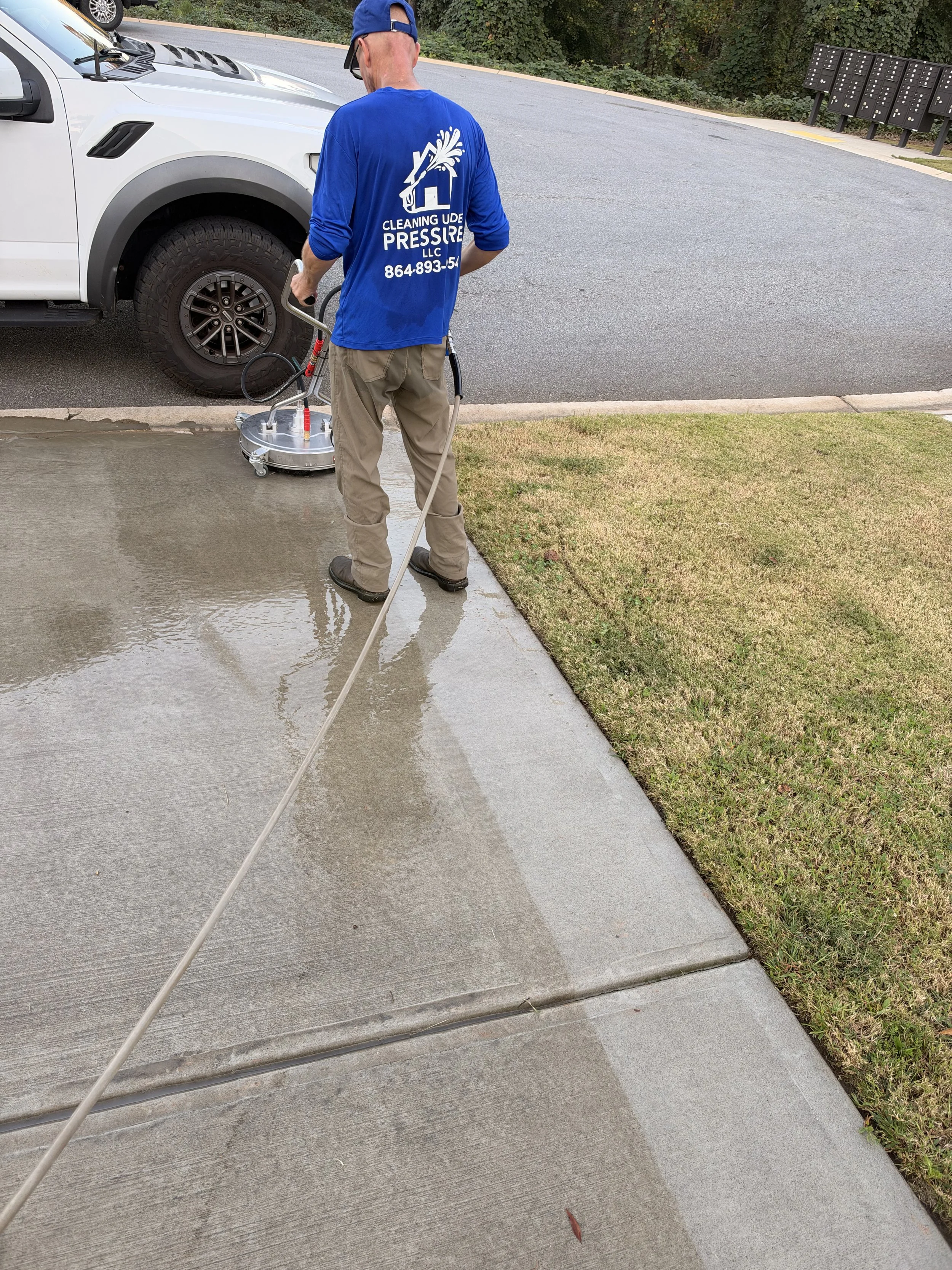 Worker pressure-washing a concrete sidewalk beside a white pickup truck