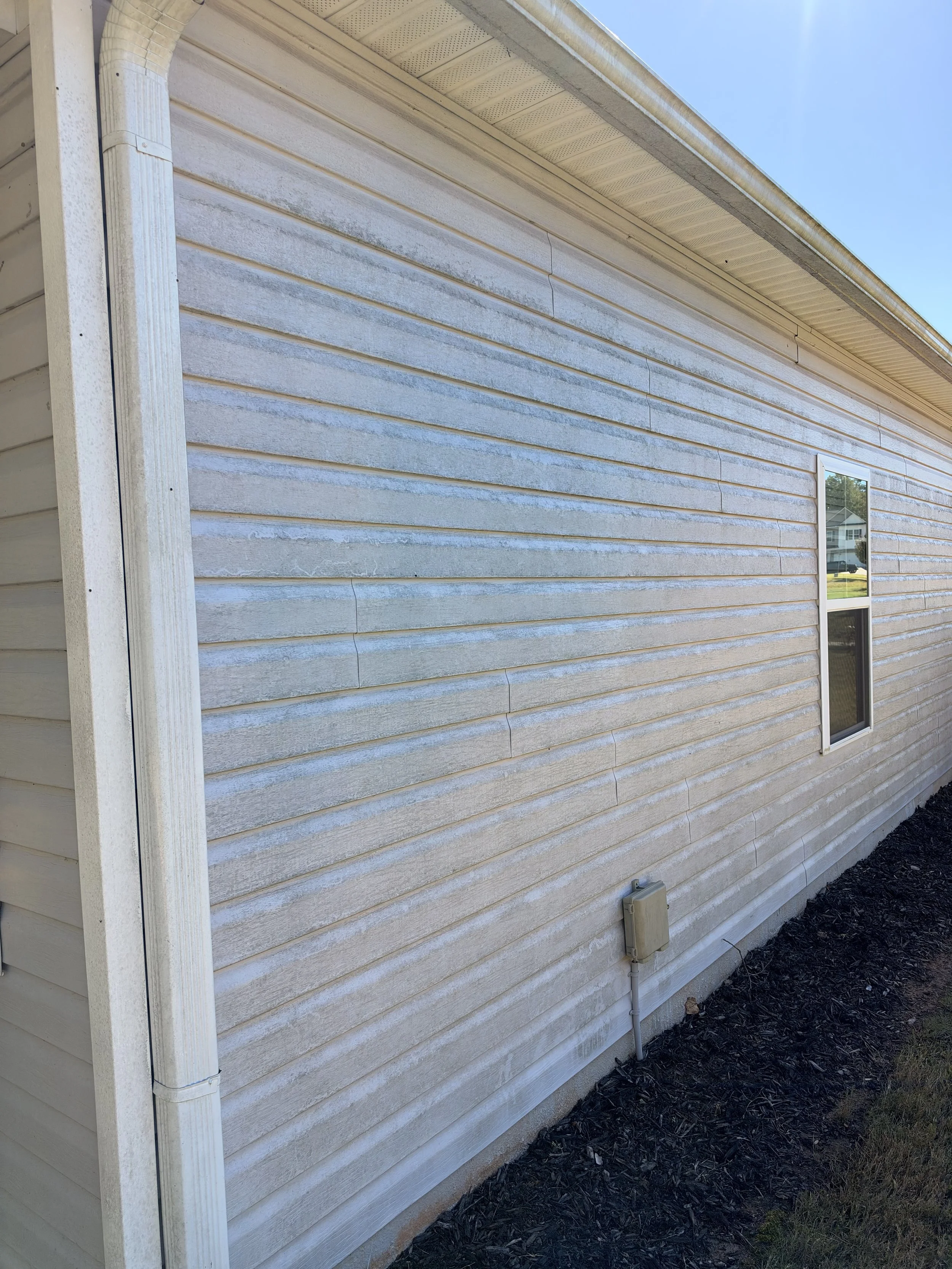 Close-up of a light-colored house exterior with horizontal siding, gutter, downspout, window, and mulched landscaping.