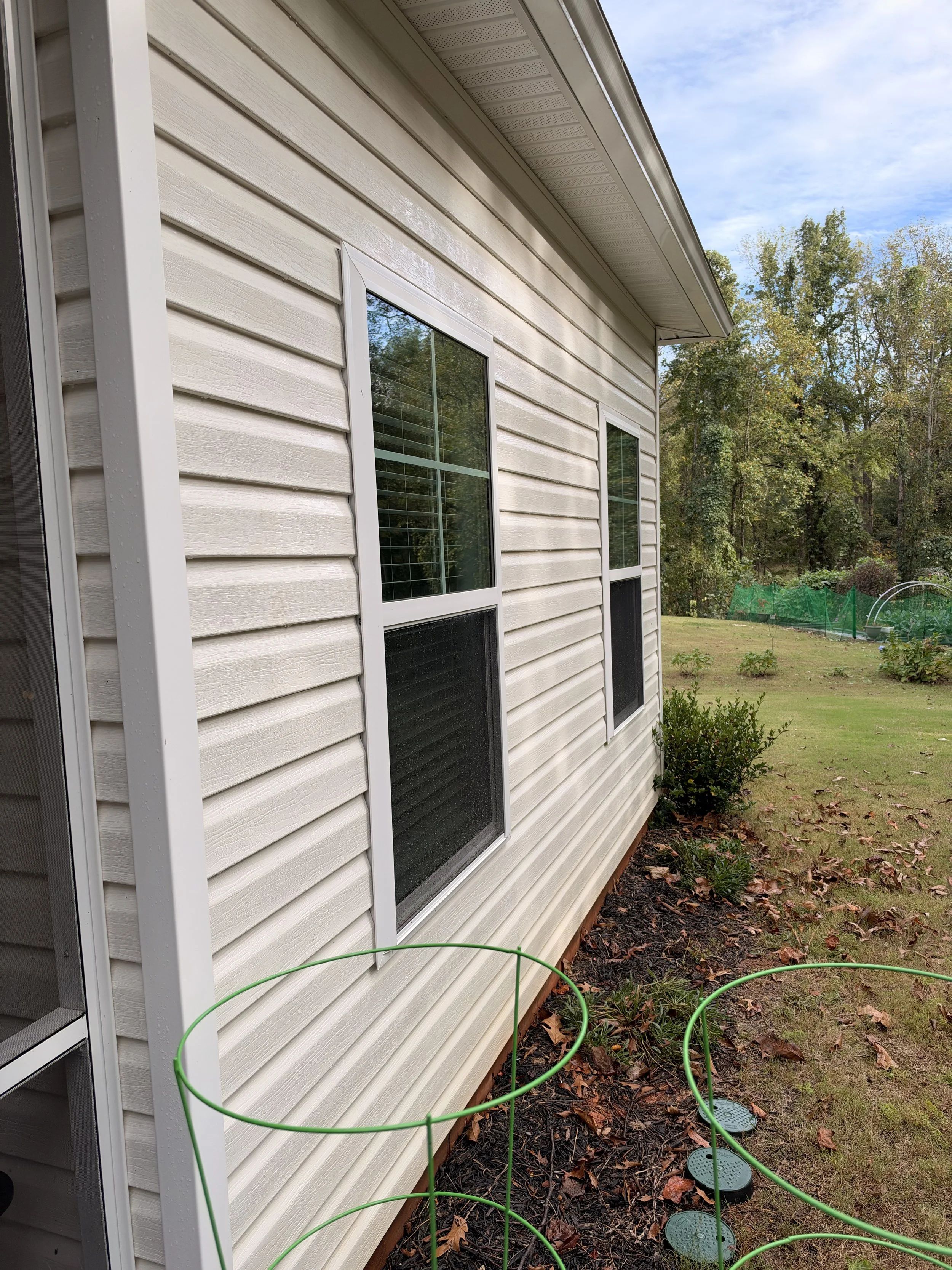 Exterior of a light-colored vinyl-sided house with screened windows, soffit, landscaped yard, garden fencing, and green garden hose.