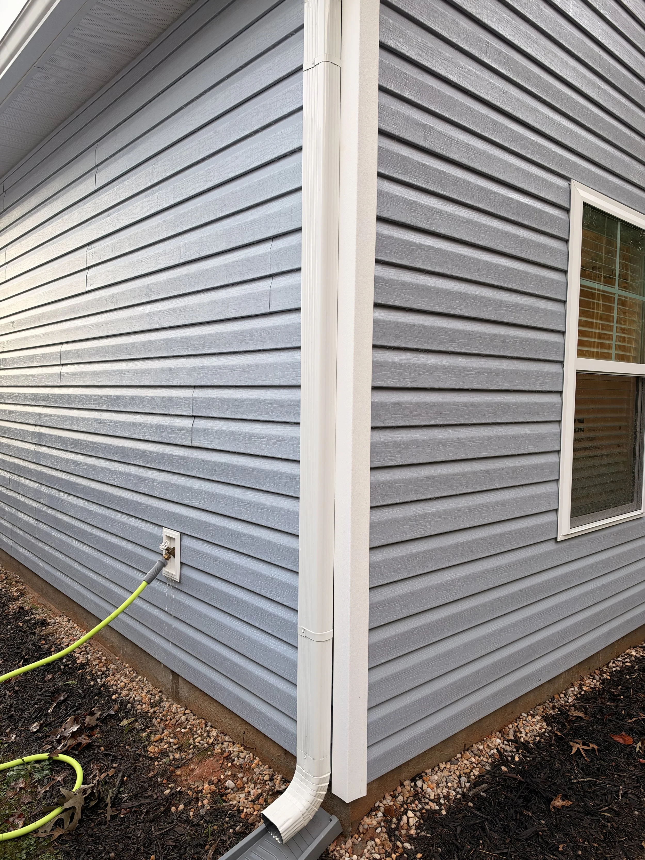 Gray vinyl-sided house exterior with white gutter downspout, window, outdoor faucet, and green garden hose.