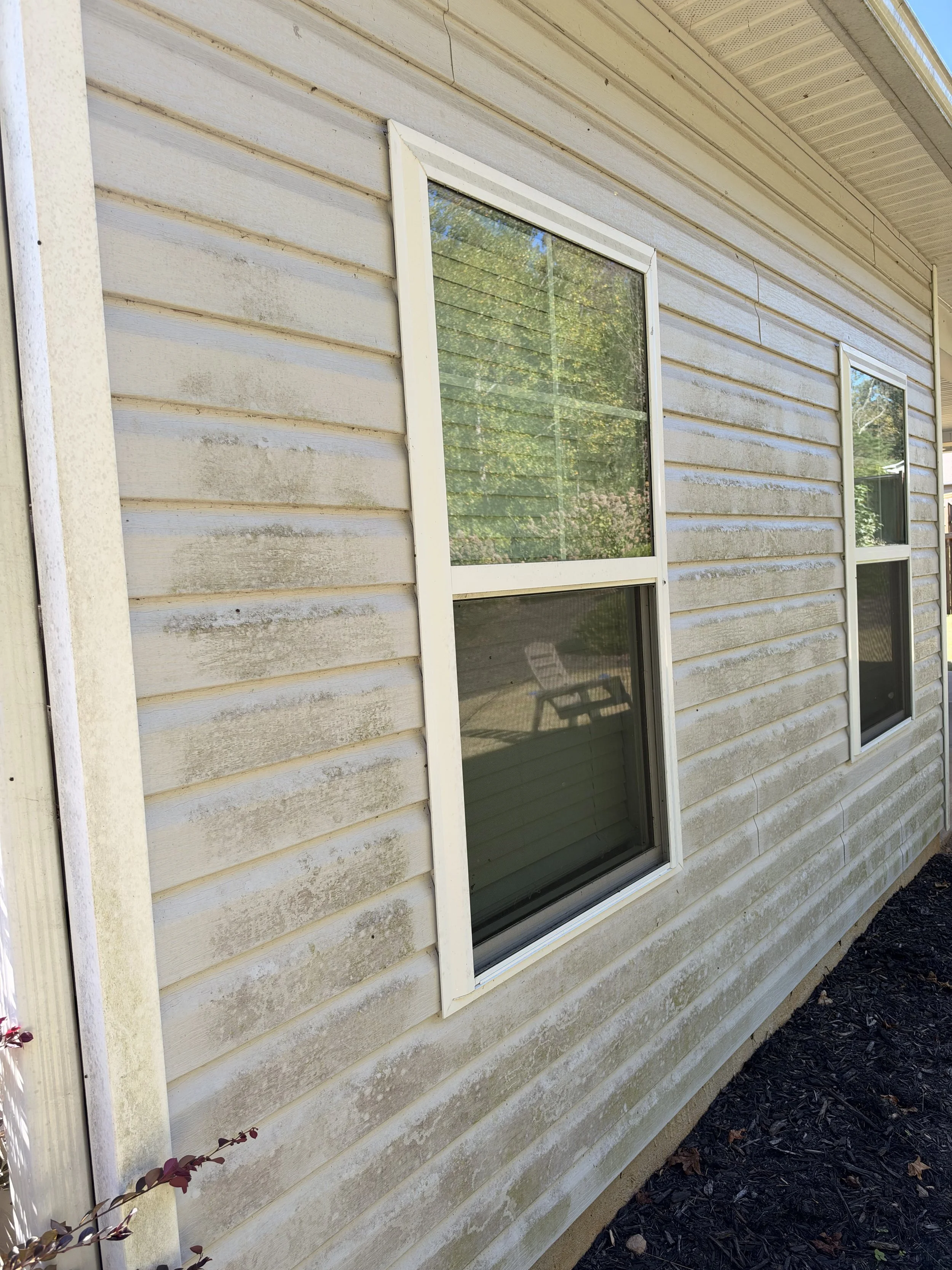 Exterior of a weathered white-sided house with two screened windows beneath a covered soffit.