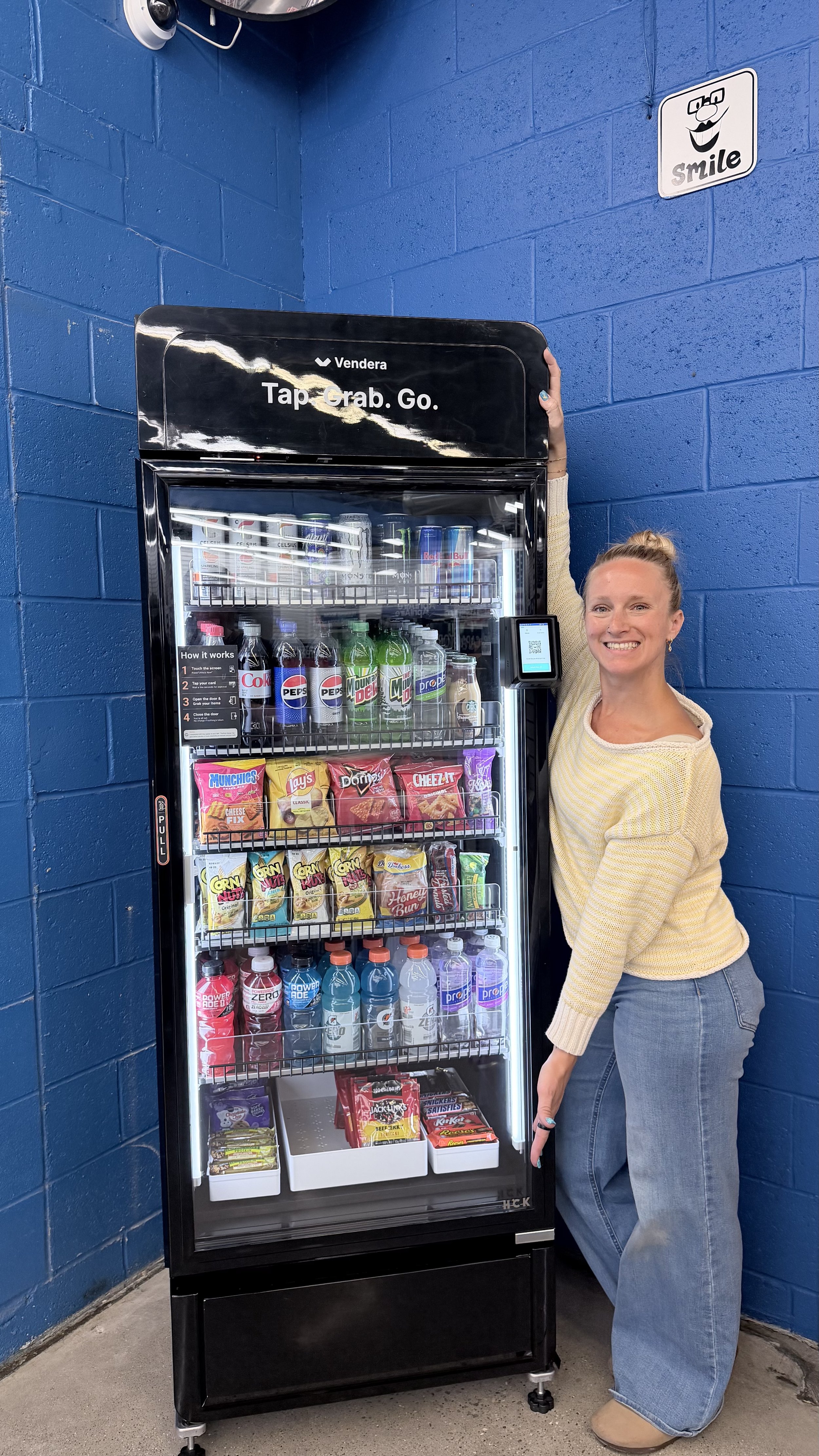 Smiling woman with blonde hair tied in a bun wearing a yellow sweater and blue jeans next to a vending machine in Dayton, Ohio against a blue brick wall.