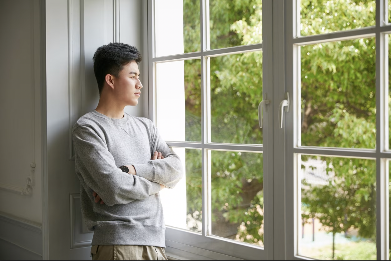 Young man in a gray sweatshirt standing with arms crossed, looking out a window at green trees outside.