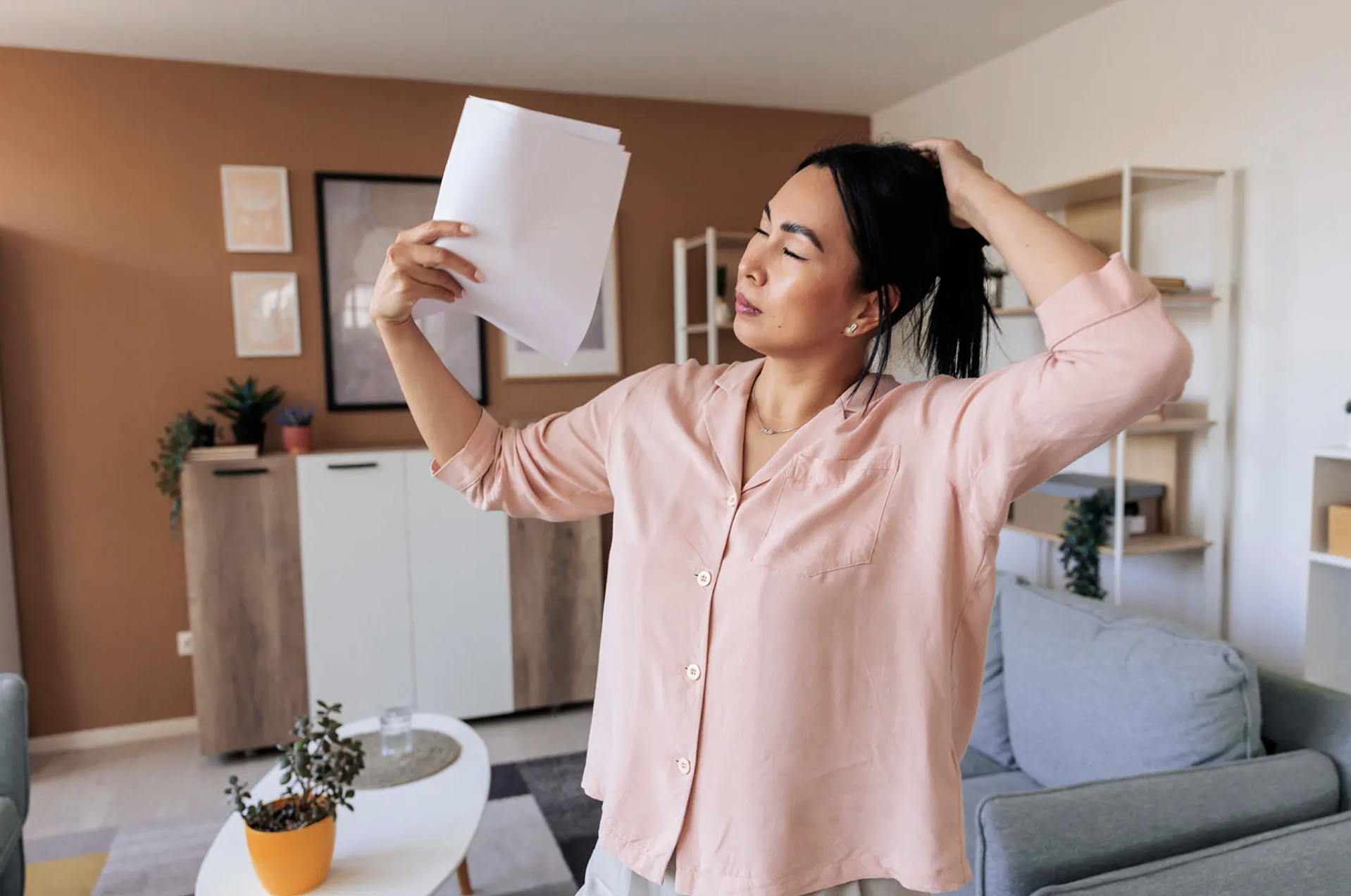A woman with dark hair and light skin is stretching indoors, holding a stack of papers in one hand and touching her head with the other, in a cozy living room.
