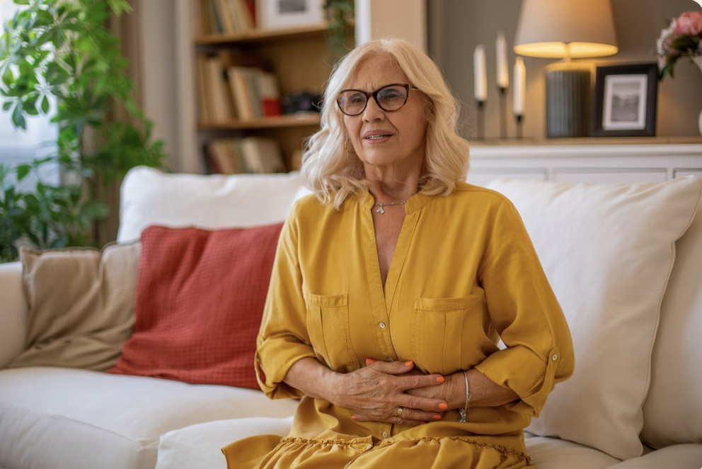 Older woman sitting on a beige couch with white and red pillows in a cozy living room. She has blonde hair, glasses, and is wearing a yellow blouse. She is holding her stomach and appears to be experiencing discomfort.