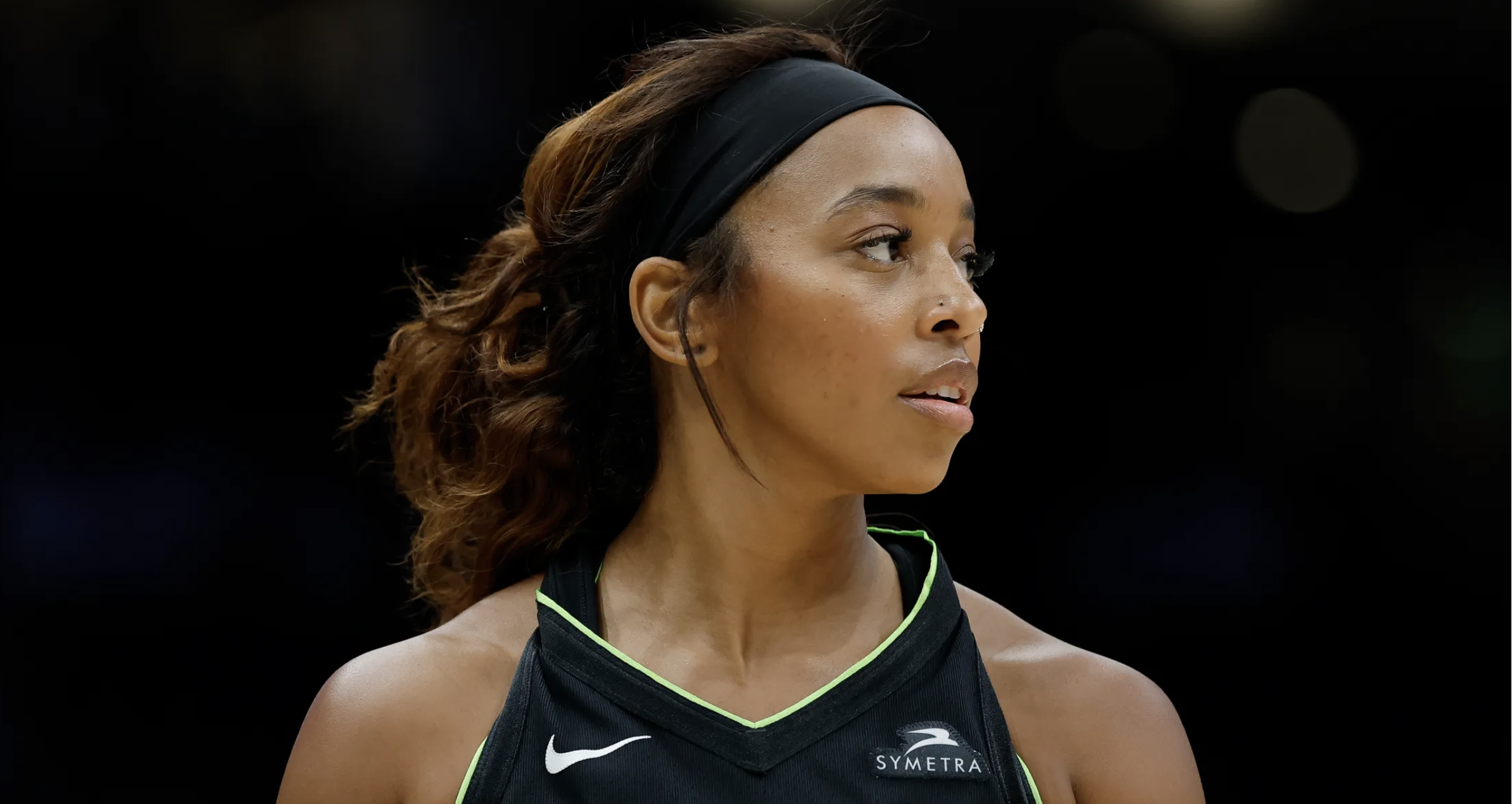 Close-up of a female athlete with curly brown hair, wearing a black sports jersey with green accents, black headband, and earrings, looking to her left with a focused expression.