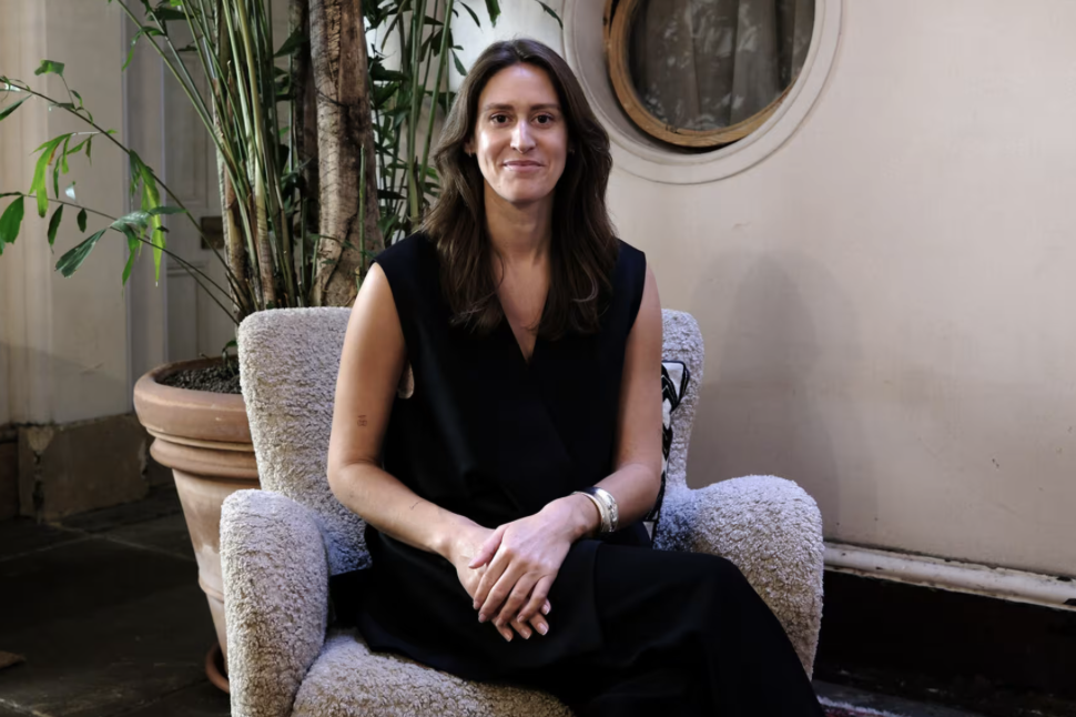 A woman with shoulder-length brown hair sitting on a gray armchair, wearing a sleeveless black dress, with a large potted plant and a round mirror on the wall behind her.