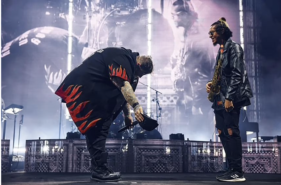 Two musicians on stage, one bowed with his head down, and the other standing and smiling, with a large LED screen behind them displaying their image.