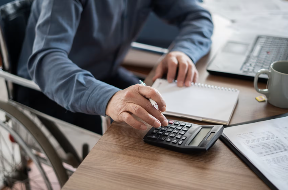 Person in a wheelchair using a calculator on a desk with notepad and documents.