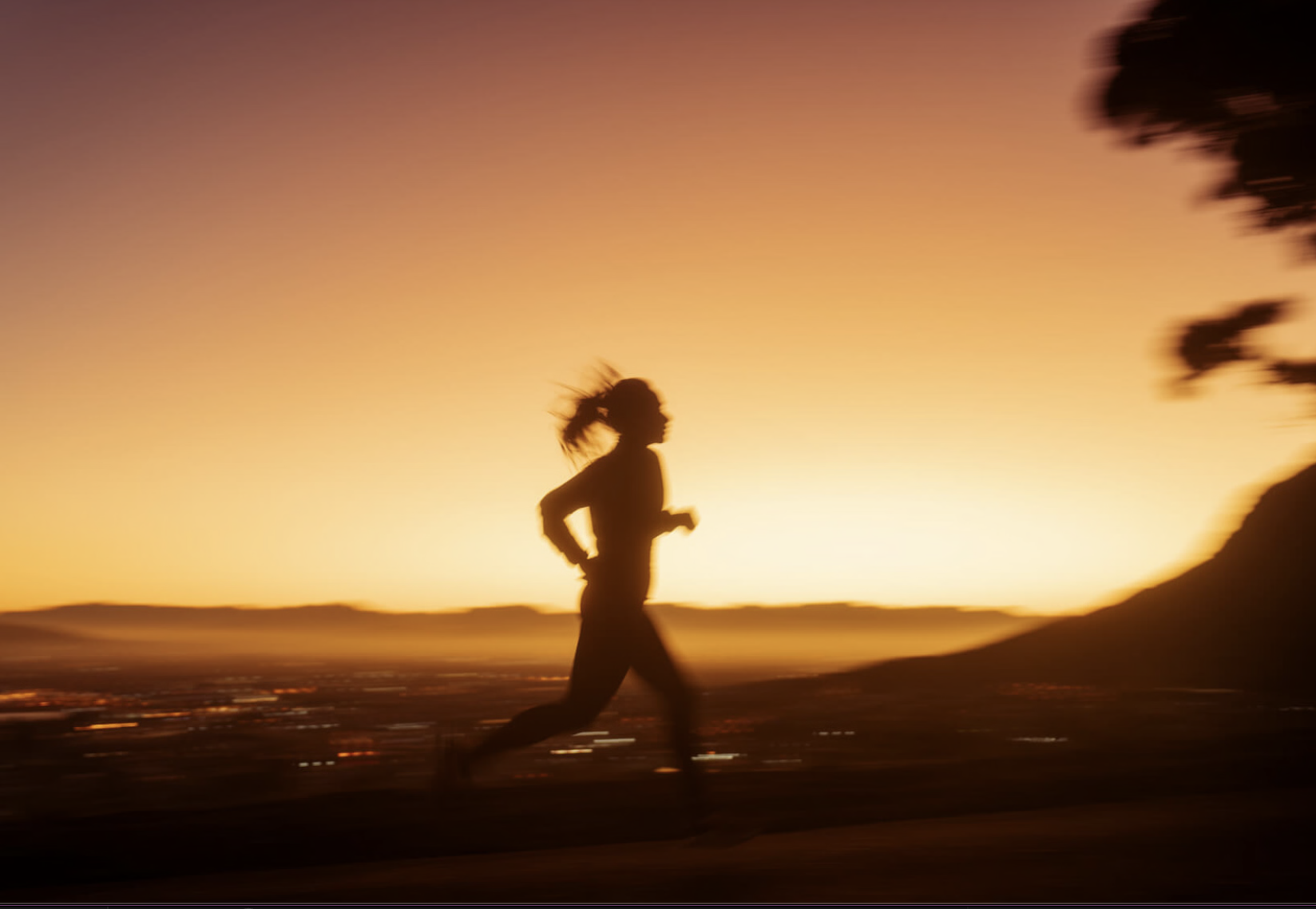 Silhouette of a person running during sunrise or sunset with a blurred cityscape and mountains in the background.