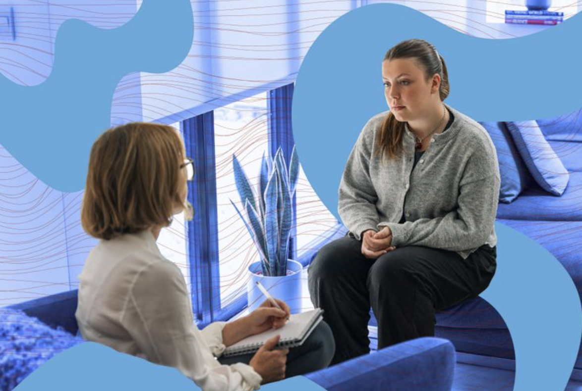 A woman in a gray sweater sitting on a blue couch, talking to a therapist who is taking notes in a notebook. The room has blue decor with a large plant in a pot and patterned wallpaper.