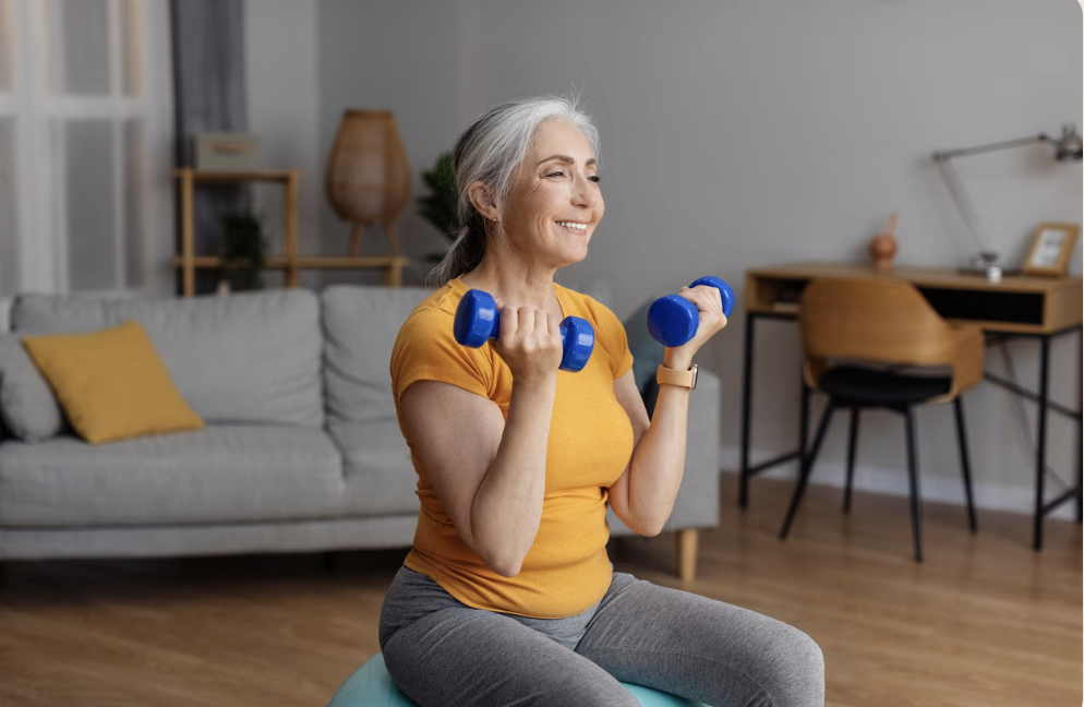 An elderly woman with gray hair, wearing a yellow shirt and gray pants, is exercising with blue dumbbells in a living room. She is smiling and sitting on a workout ball.