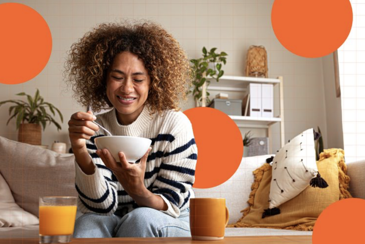 A woman with curly hair sitting on a couch, smiling, holding a bowl and spoon, with a glass of orange juice and a mug on the table in front of her, in a cozy living room with plants and pillows.