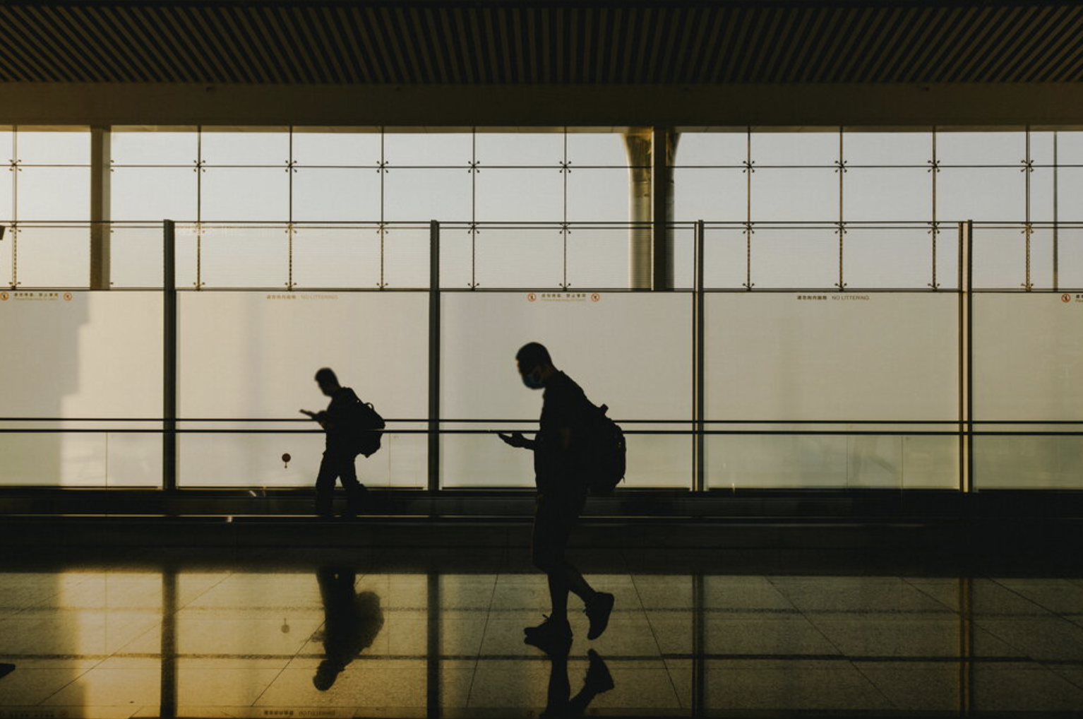 Silhouettes of two people wearing face masks and backpacks walking in an airport terminal, looking at their phones. The background features large windows with a grid pattern and a yellowish light coming through.