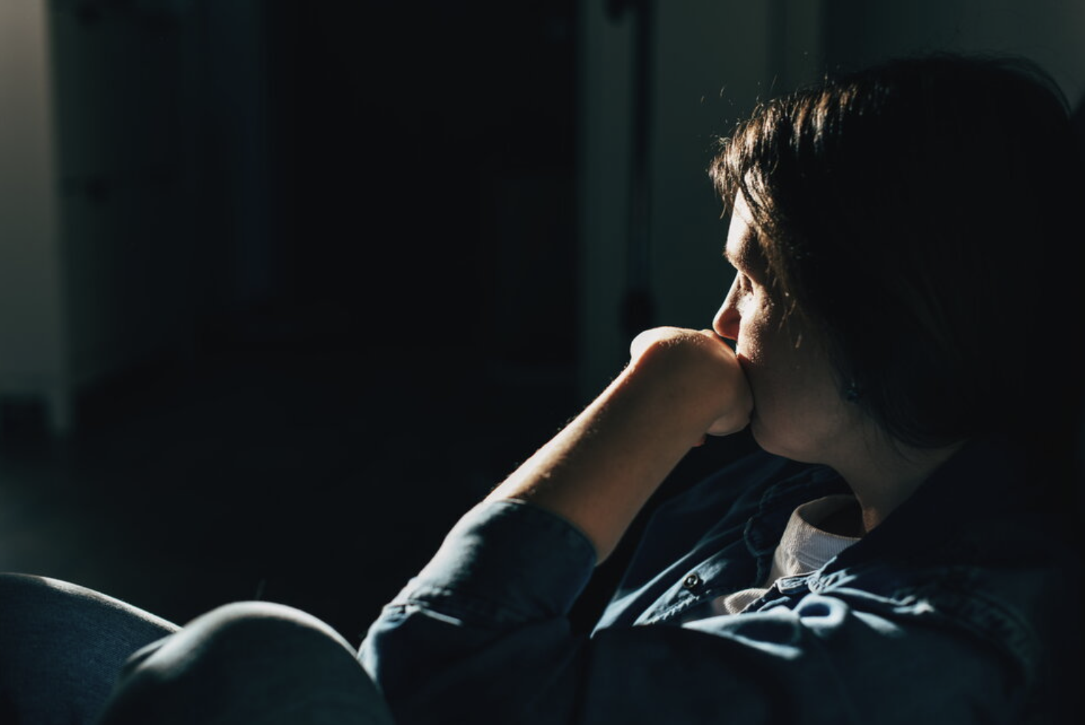 A woman sitting in a dark room, resting her head on her knee, with sunlight on her face.