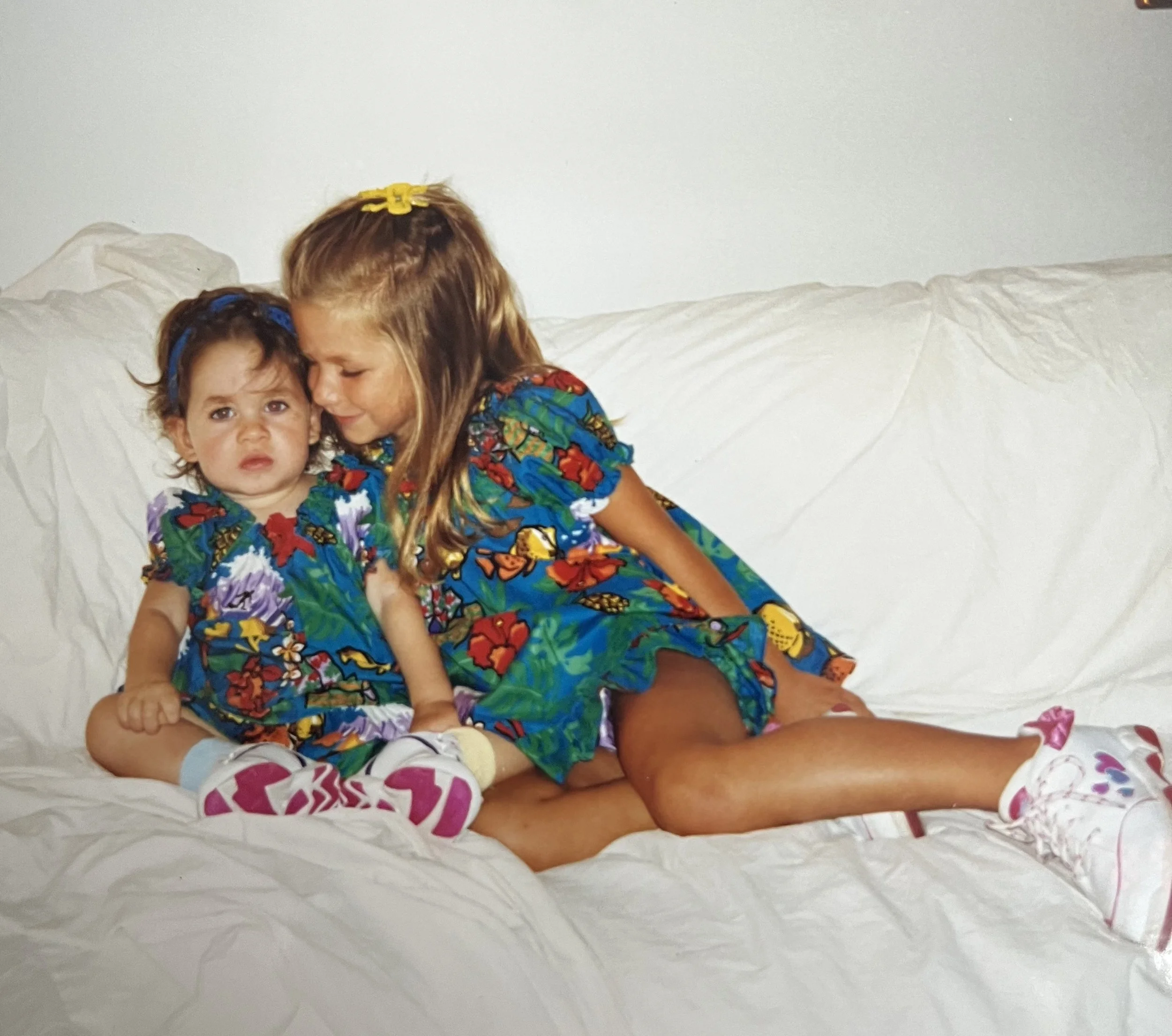 A young woman and a small girl sitting on a bed, close together, with a plain white wall in the background. The girl looks directly at the camera with a slightly confused expression, while the woman leans in with her eyes closed, appearing to whisper or kiss the girl's temple. Both are wearing colorful, patterned clothing and have hair clips in their hair.