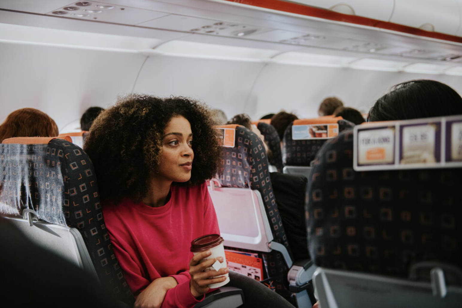 A woman with curly hair, wearing a pink sweater, sitting in an airplane seat, holding a coffee cup, looking towards the window.