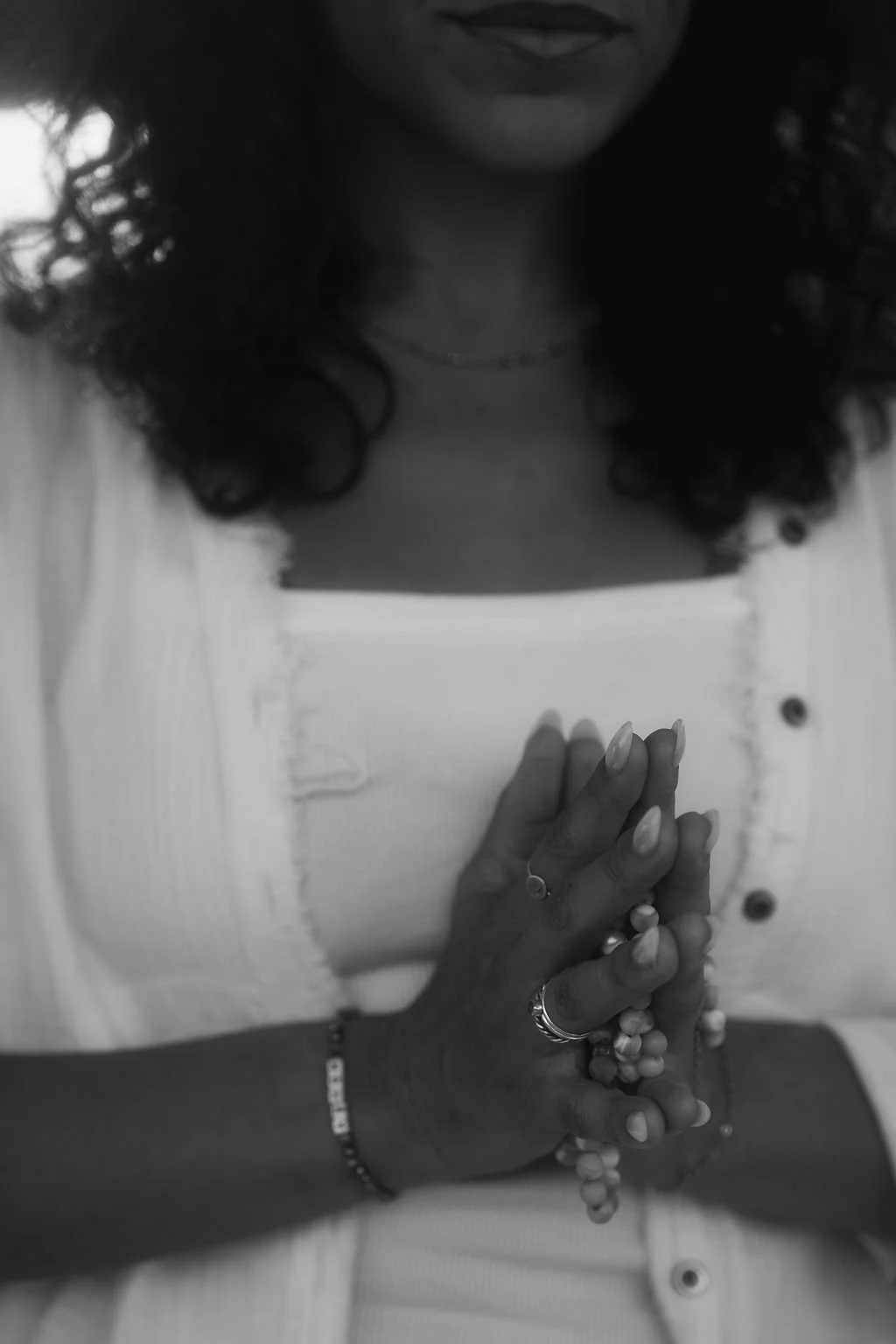 A woman with curly hair and light skin holding prayer beads with both hands in a meditative pose, wearing a white knitted cardigan over a white top, and a necklace.