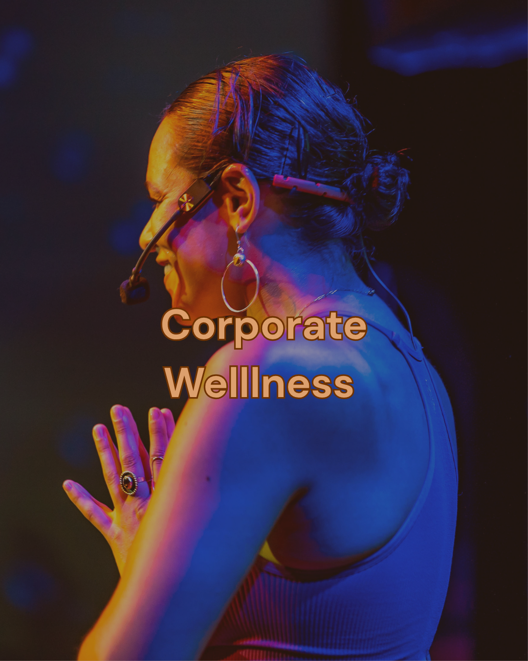 A woman with bun hairstyle, wearing glasses and hoop earrings, seen from the side with her hands pressed together in a prayer position, illuminated with colorful stage lights. Overlaid text reads 'Corporate Wellness'.