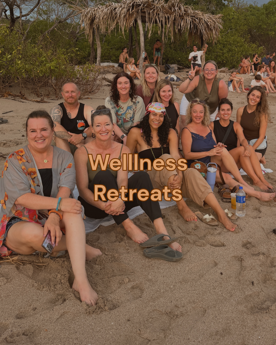 Group of people sitting and standing on the beach under a thatched roof with trees in the background, participating in a wellness retreat.