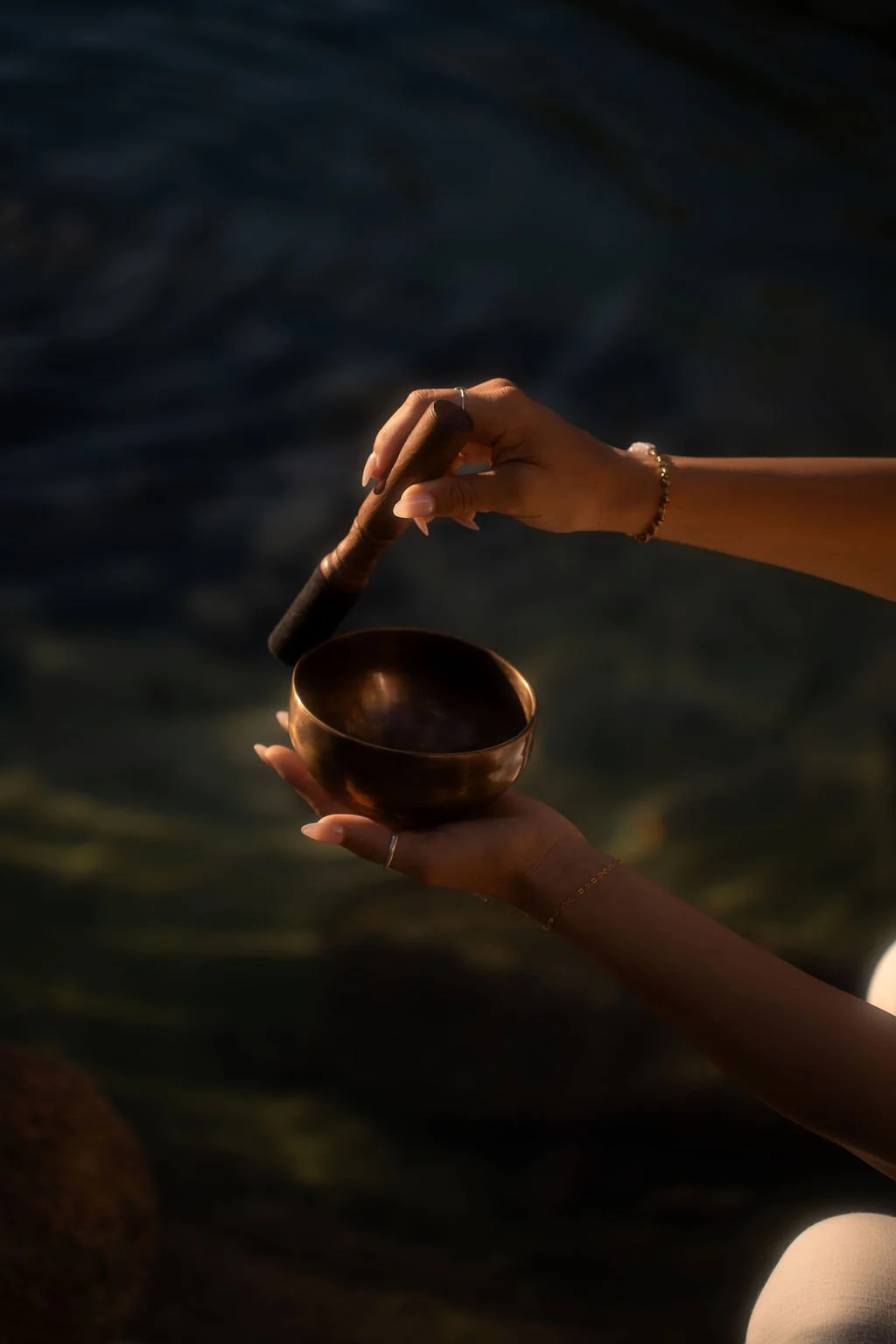 Close-up of a person holding a singing bowl in their left hand and a wooden mallet in their right hand, with a dark water background.