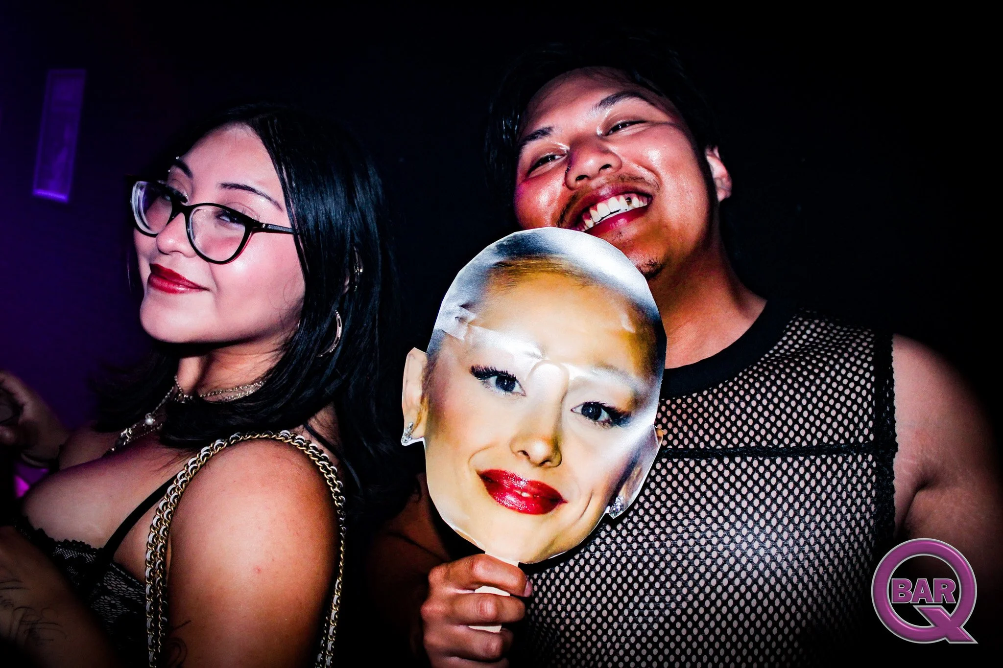 Three people in a dark setting, with two of them holding a face mask with a woman's face on it. The woman on the left has long black hair, glasses, and is dressed in black. The man on the right has short black hair, a mustache, and is smiling. The fa