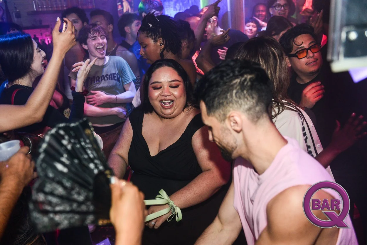 People dancing and enjoying themselves at a lively party or nightclub with colorful lighting. A woman in a black dress appears to be singing or laughing, while others around her are smiling, dancing, and having fun.