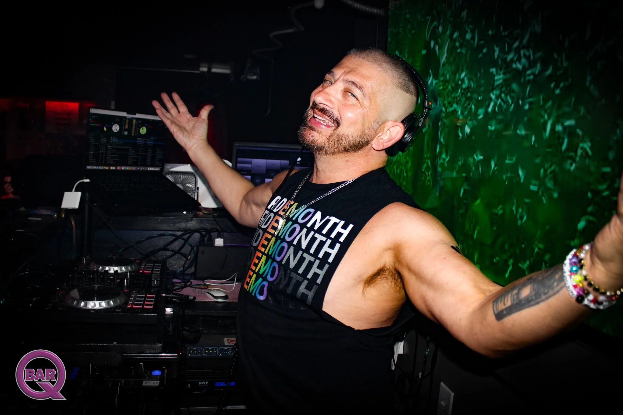 A DJ with a beard, tattoos, and short hair smiling with arms outstretched, wearing headphones and a black tank top, standing behind DJ equipment in a nightclub with green lighting and a wall of greenery.