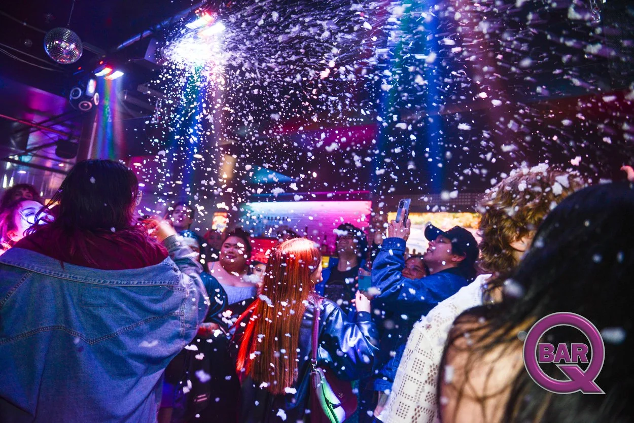 People dancing and taking photos in a nightclub with colorful lights and foam spray, including a woman with red hair and a man in a baseball cap, during a lively party.