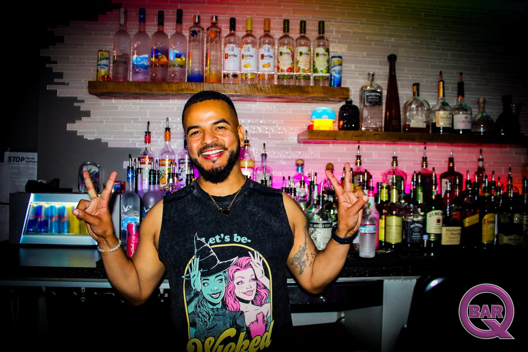 A smiling man with tattoos on his arms and a beard, posing at a bar with a peace sign with both hands. The bar has shelves filled with various liquor bottles and mixers, and the background features a colorful, illuminated display.