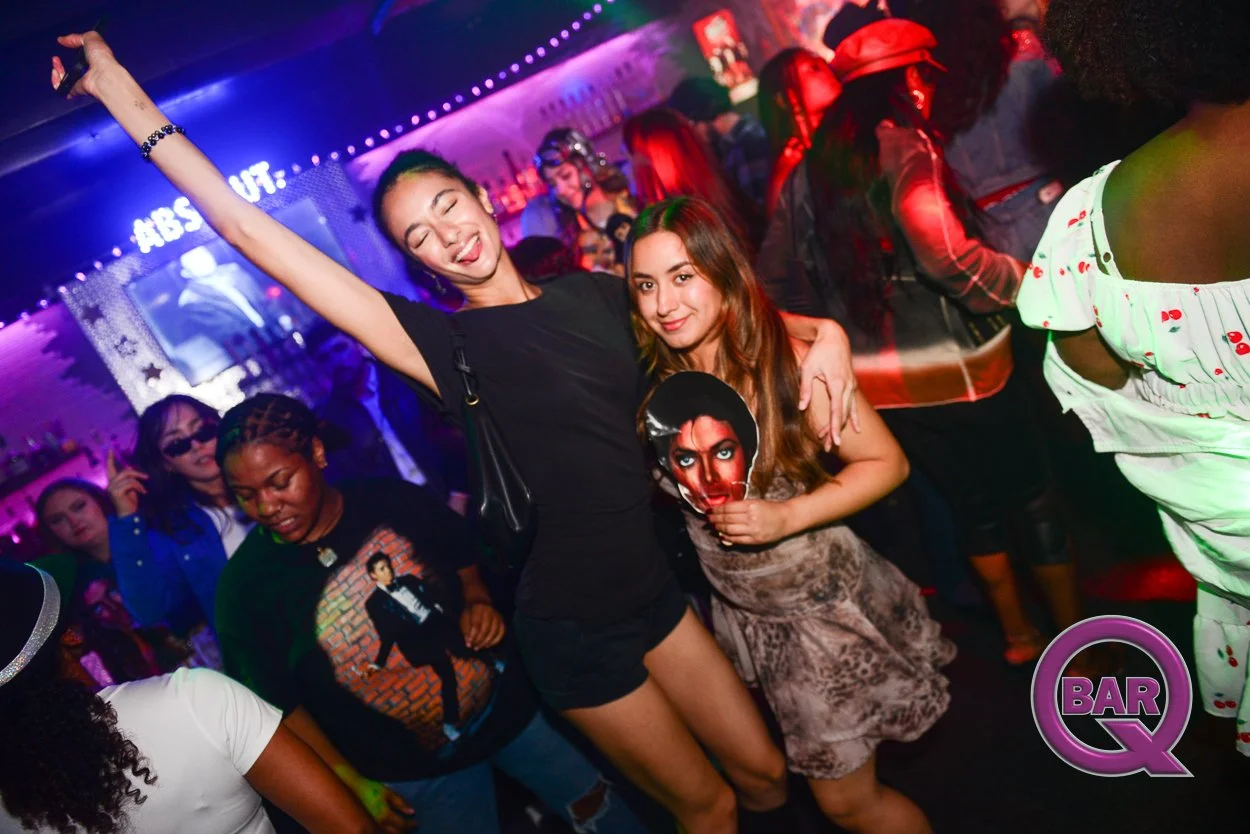 People dancing and having fun at a nightclub with colorful lighting, including a woman in a black dress and another woman holding a Michael Jackson poster.