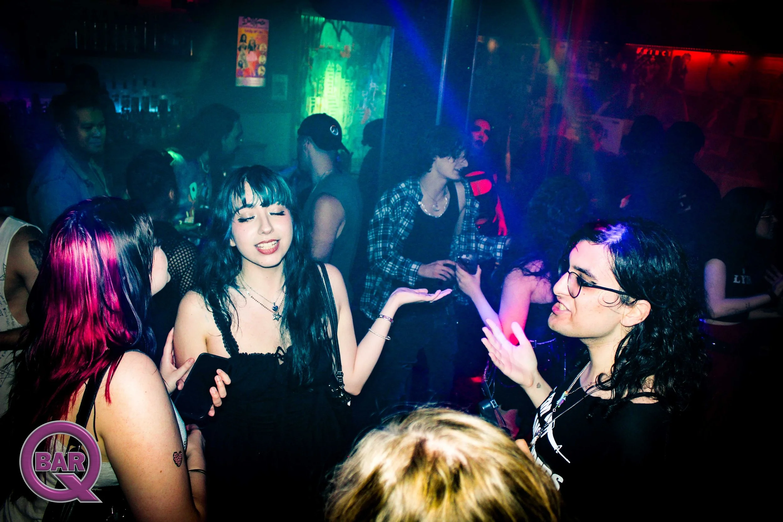 People dancing and socializing in a dark, colorful nightclub with vibrant lighting and a bar area in the background.