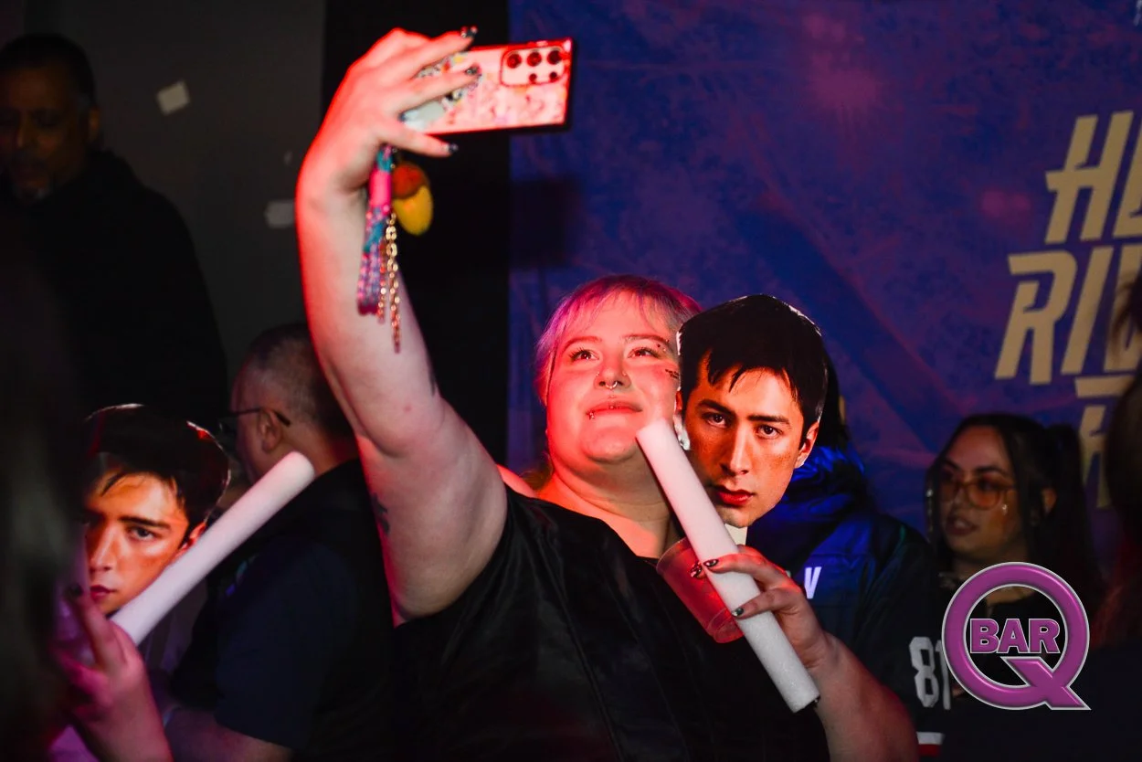 People at a party taking a selfie, with one woman holding a phone and a drink, surrounded by others in a lively atmosphere.